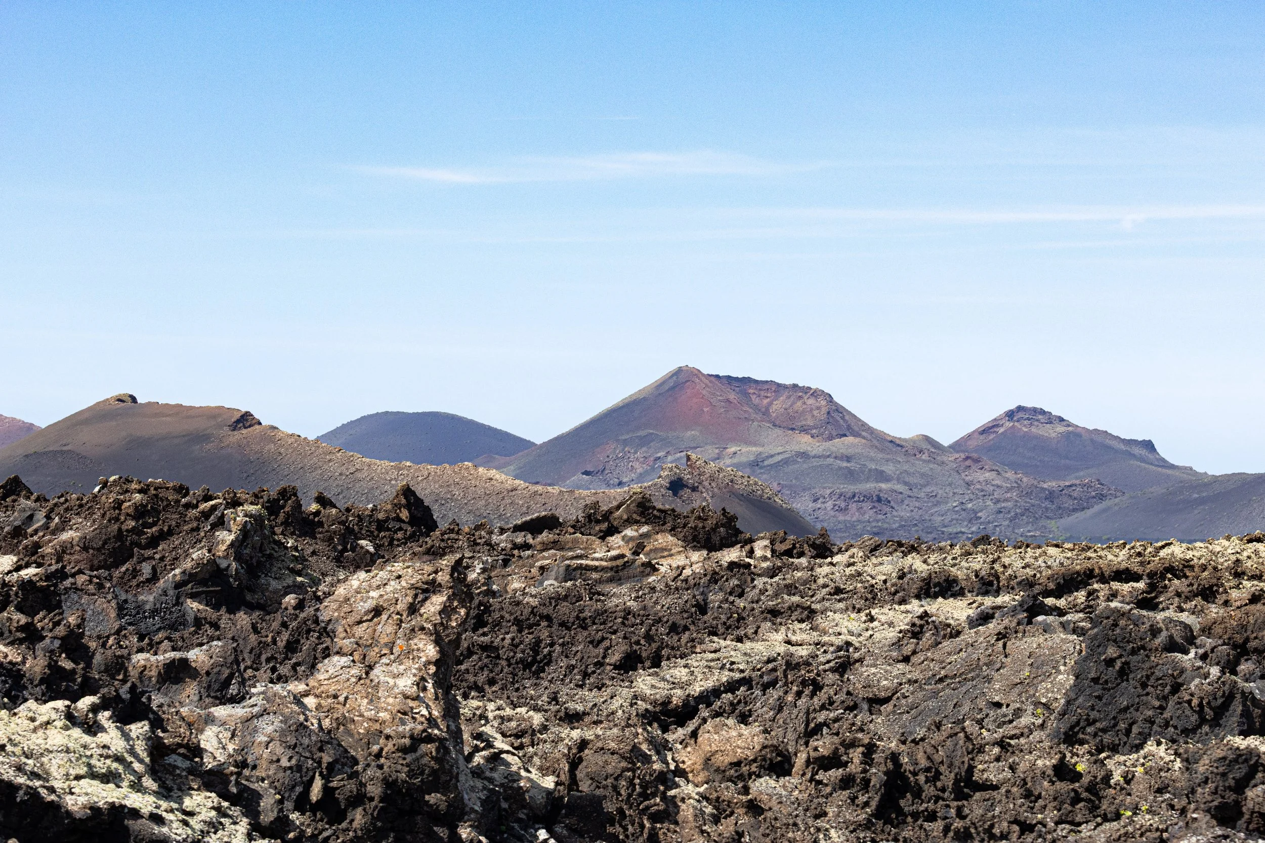 Volcanic landscape with dark, jagged lava rocks in the foreground and distant, colorful volcanic mountains under a blue sky.