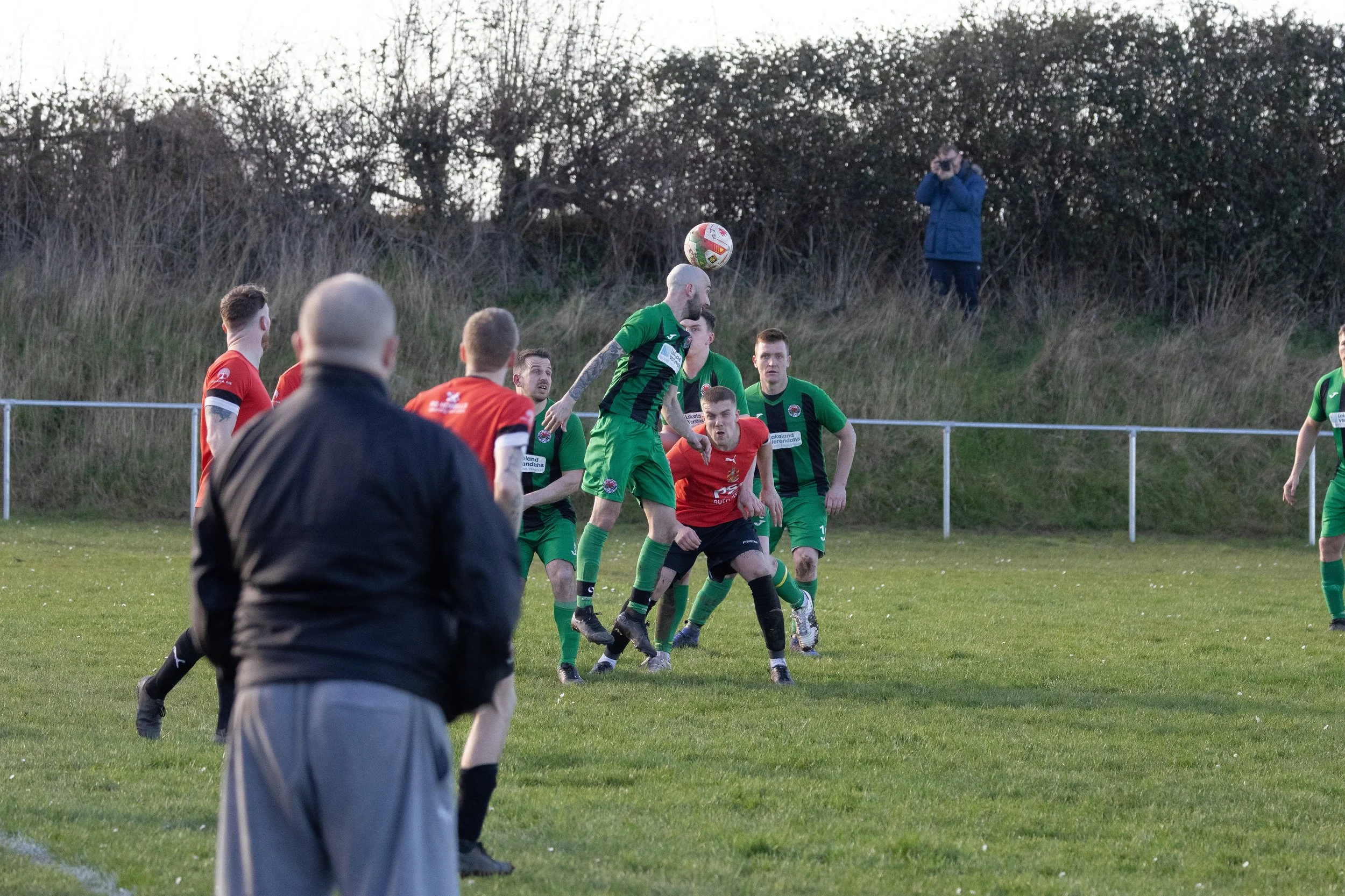 Soccer match in progress with players jumping for the ball; one player in green jumps higher, players in red try to block; referee in black and spectators in background.