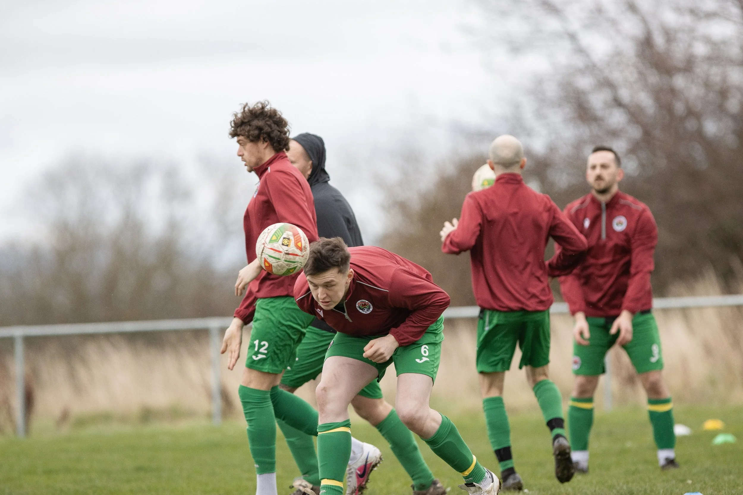 Soccer players on the field practicing with a ball, wearing green shorts and red jackets, during daytime.