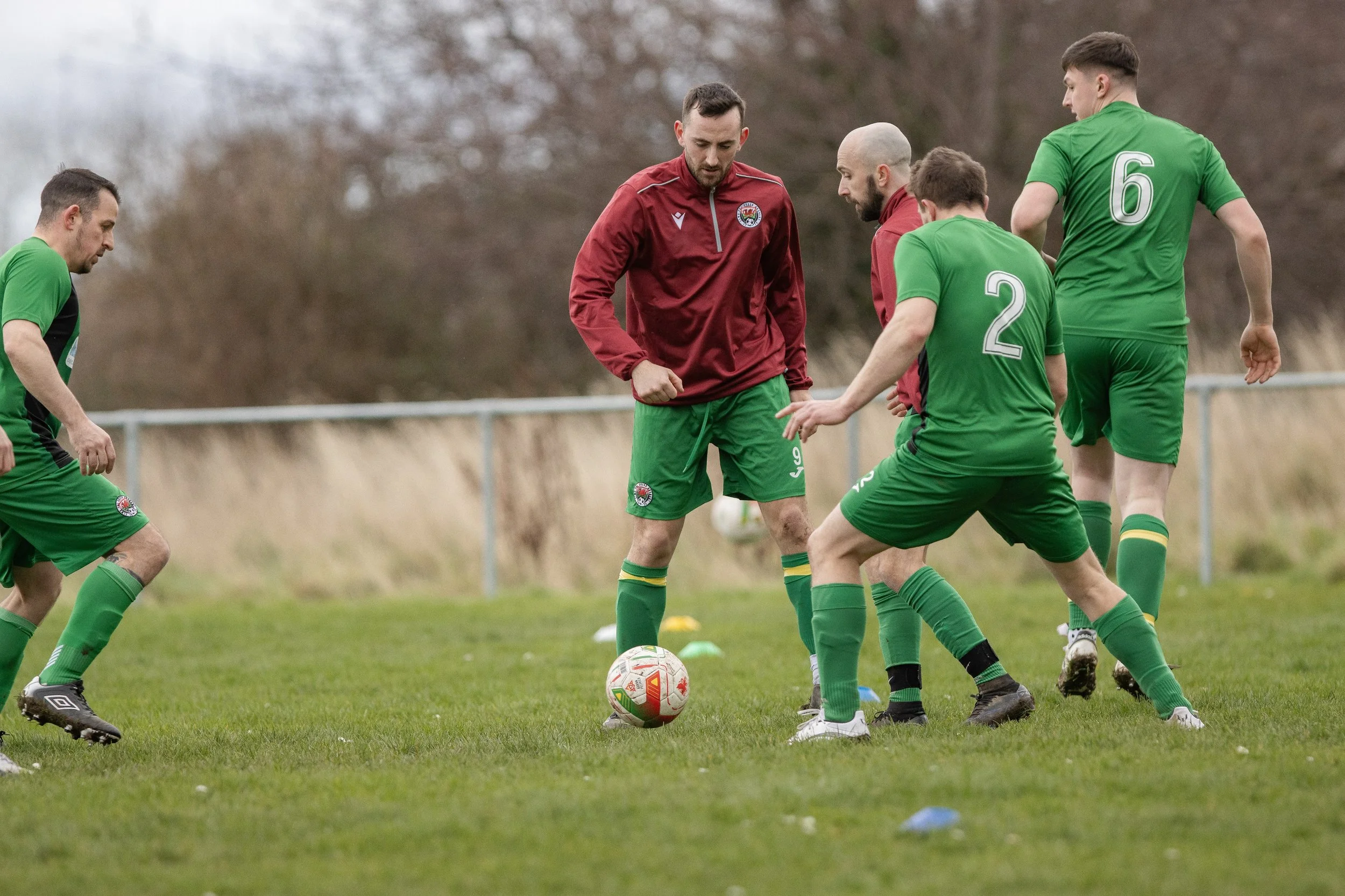 Soccer players in green uniforms practicing on a field, with one player in a red jacket preparing to kick the ball.