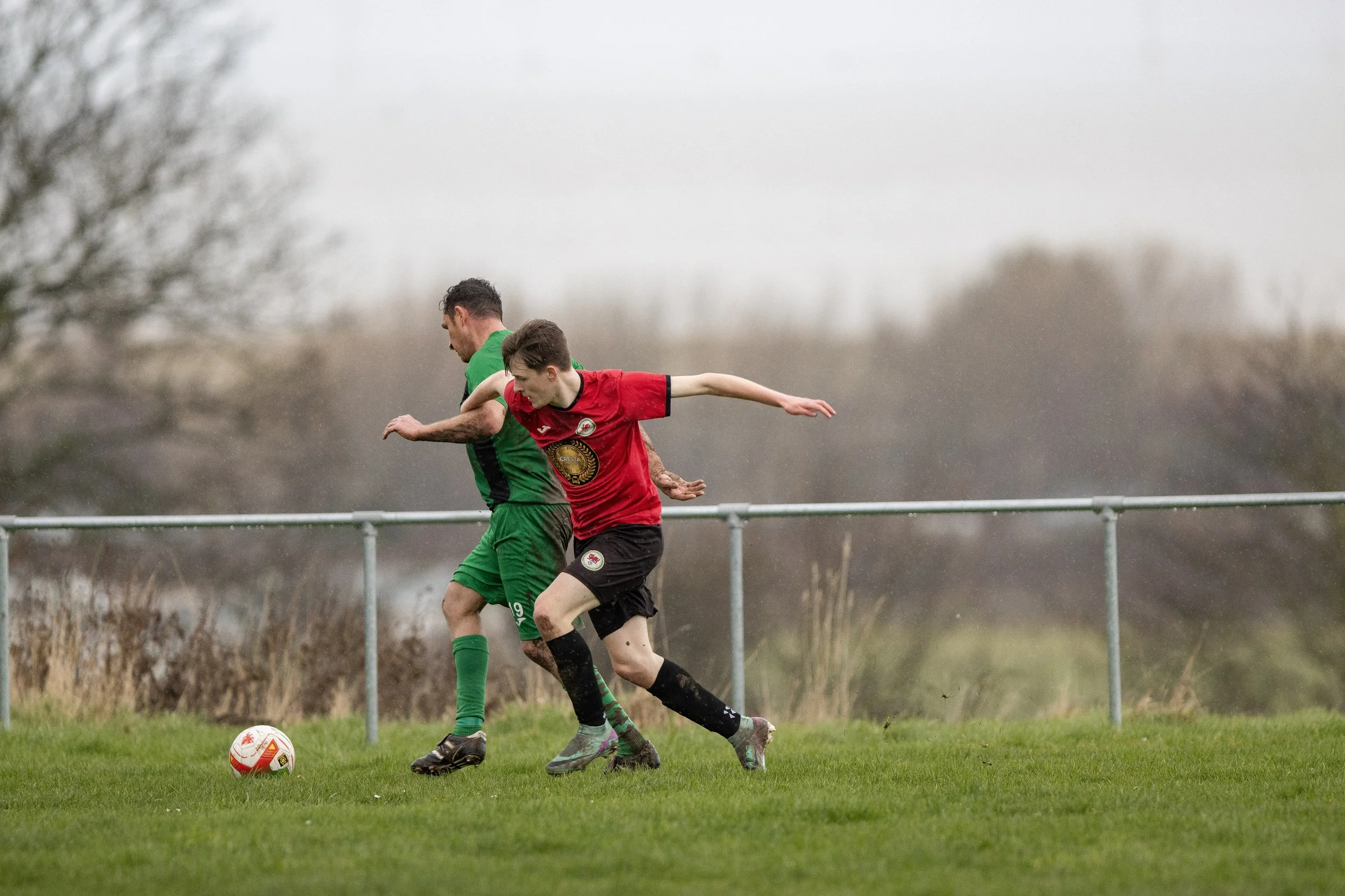 Two soccer players in green and red jerseys compete for the ball on a grassy field under a cloudy sky.