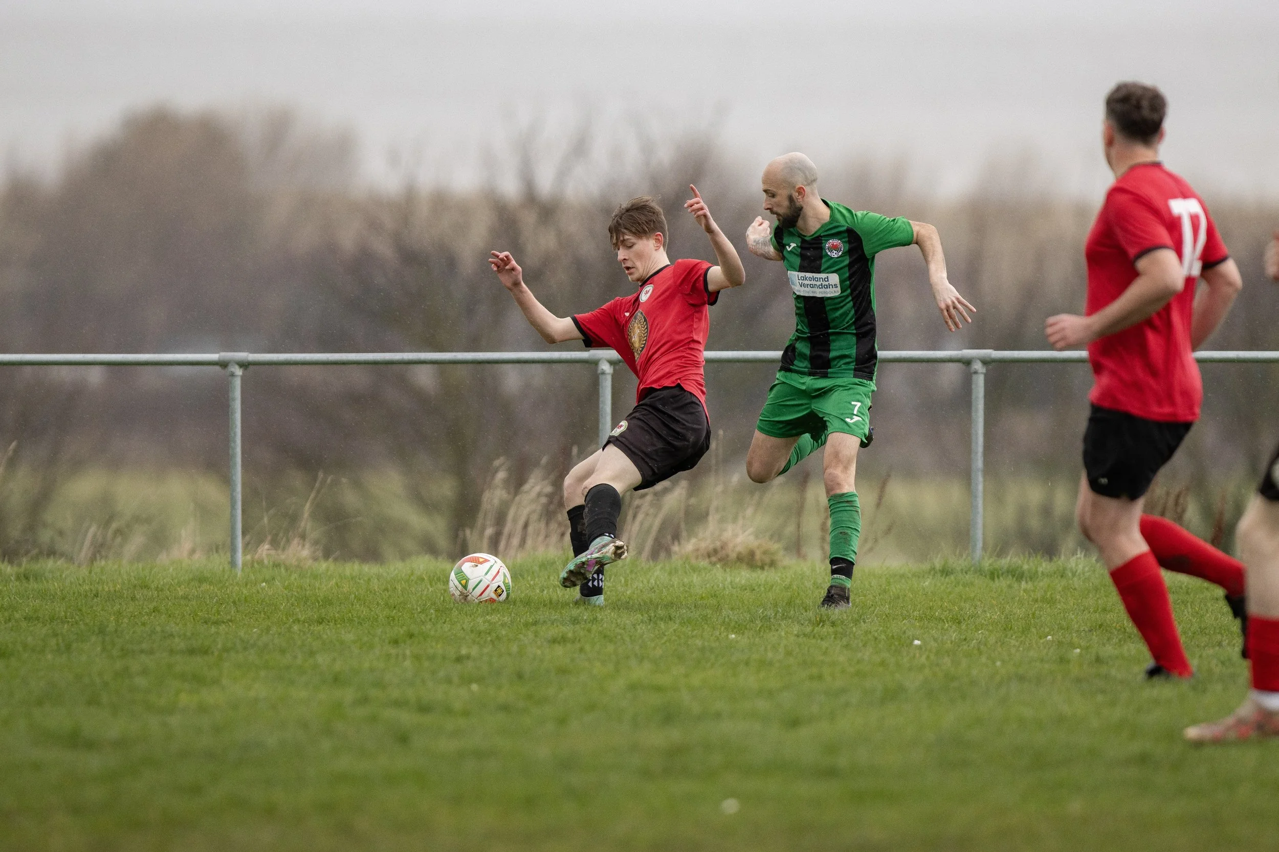 Two soccer players in red and green uniforms competing for the ball on a grassy field, with two other players in red nearby, during an outdoor match on a cloudy day.