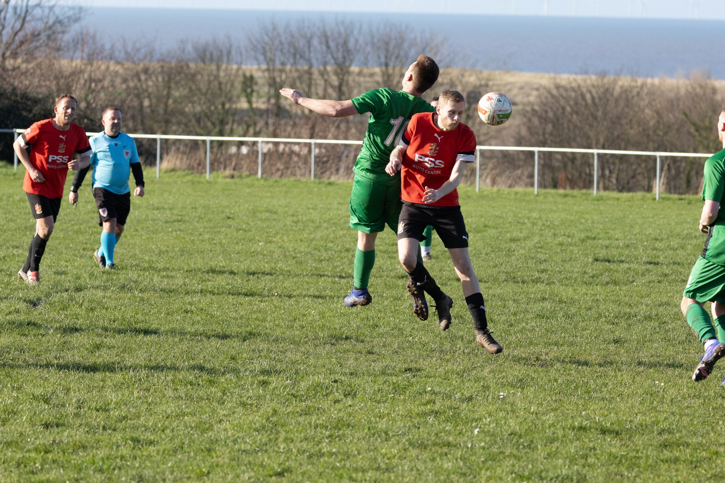 Soccer players in red and green uniforms jumping to head the ball during a match on a grassy field, with additional players and a scenic landscape in the background.