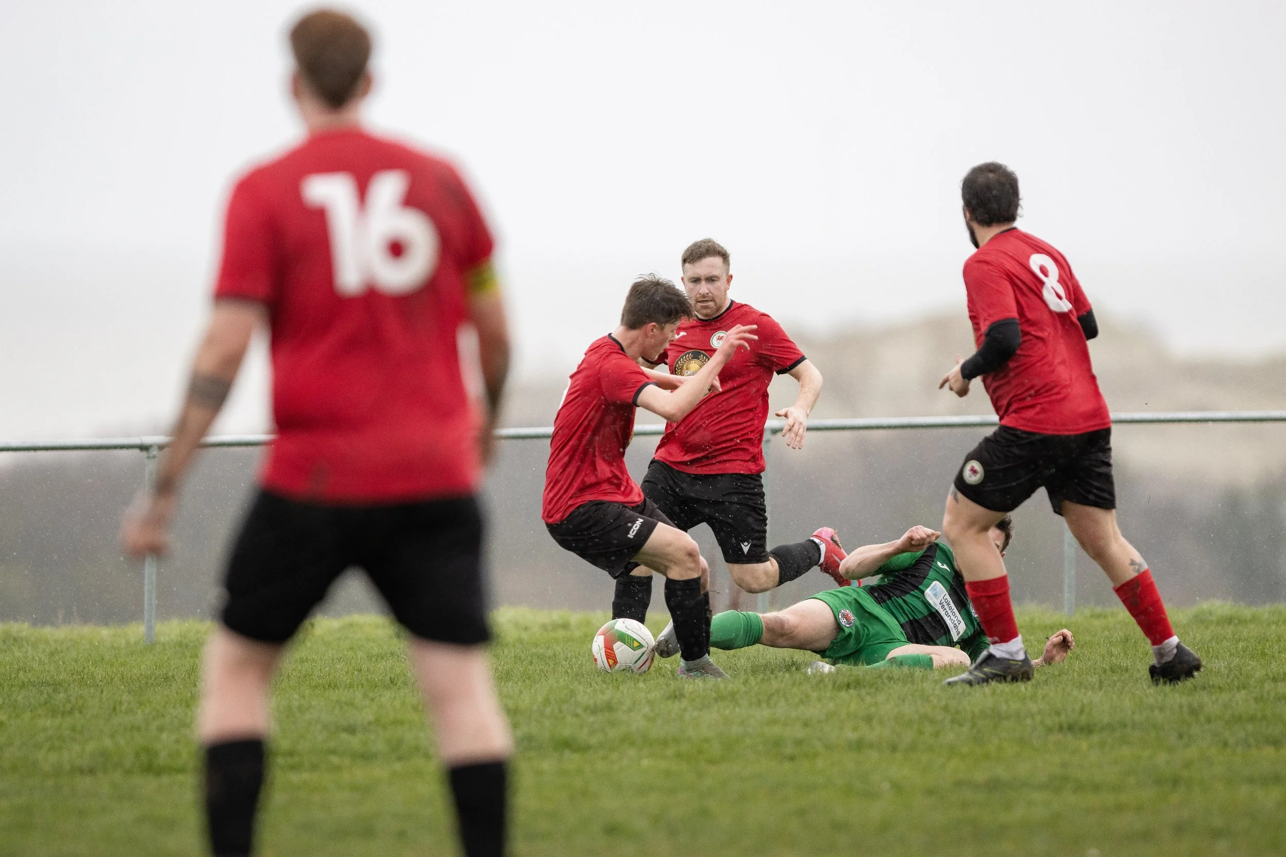 Soccer players in red jerseys and black shorts competing for the ball, with one player in a green goalkeeper uniform on the ground.