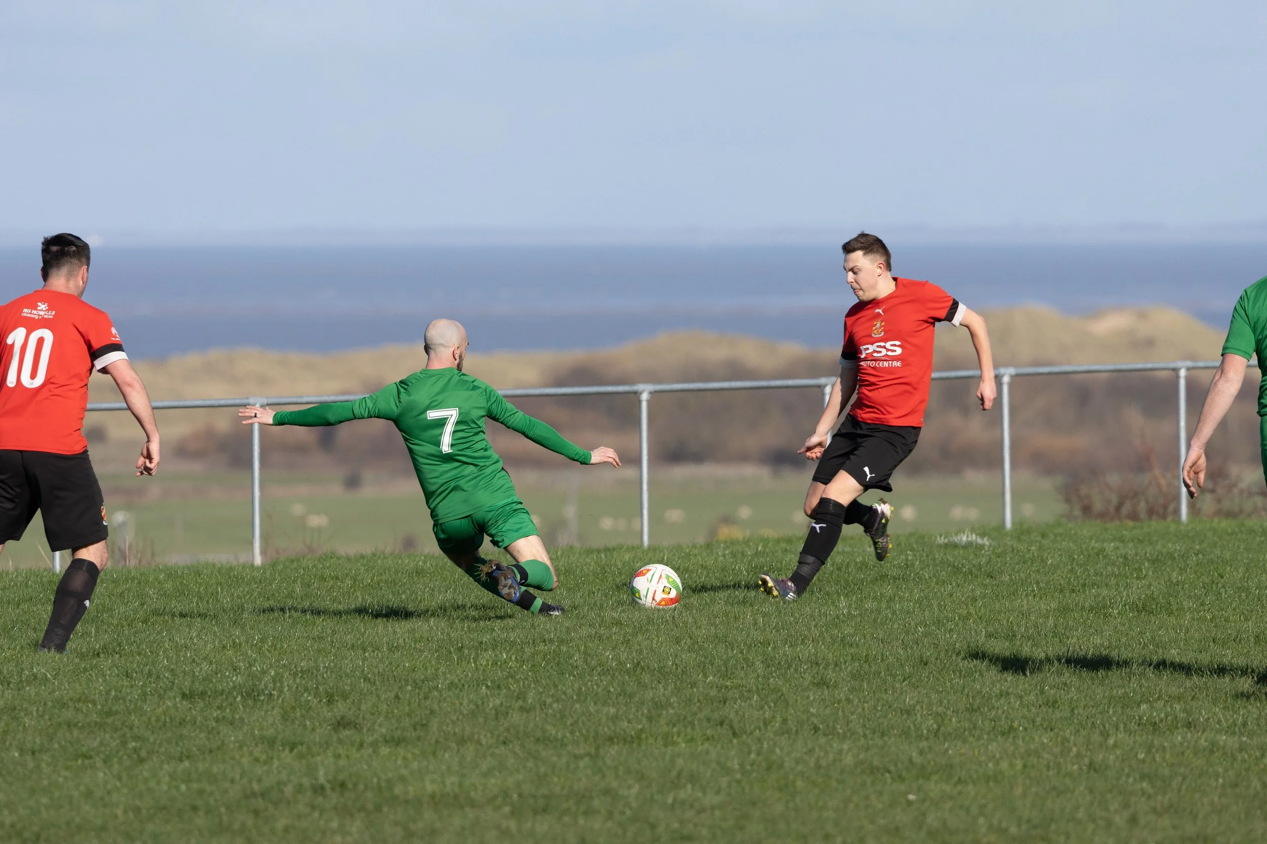 Soccer players on the field during a match, with the goalkeeper in green attempting to block or save the ball from an opposing player in a red jersey.