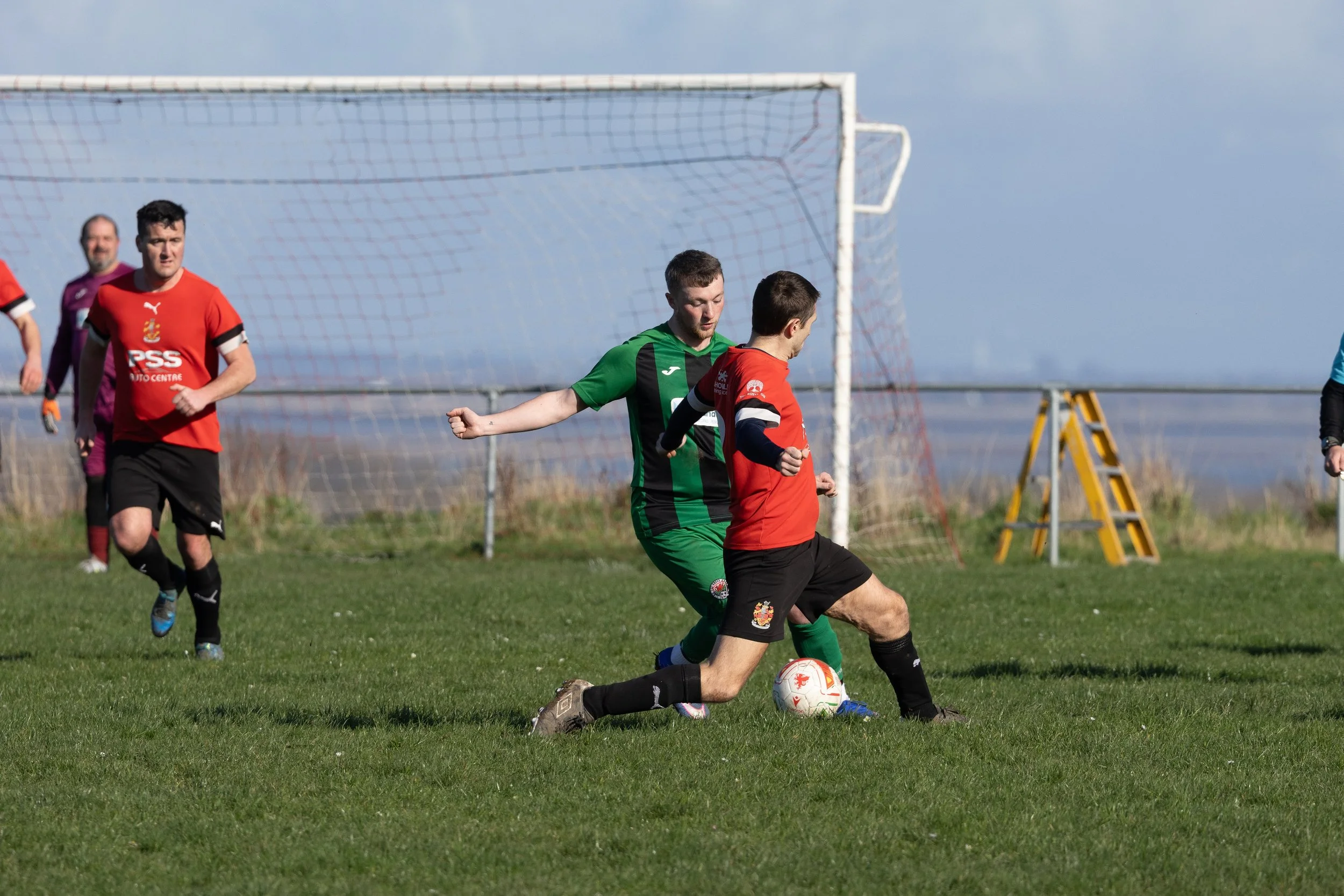 Soccer players competing for the ball on the field, with a goal in the background.
