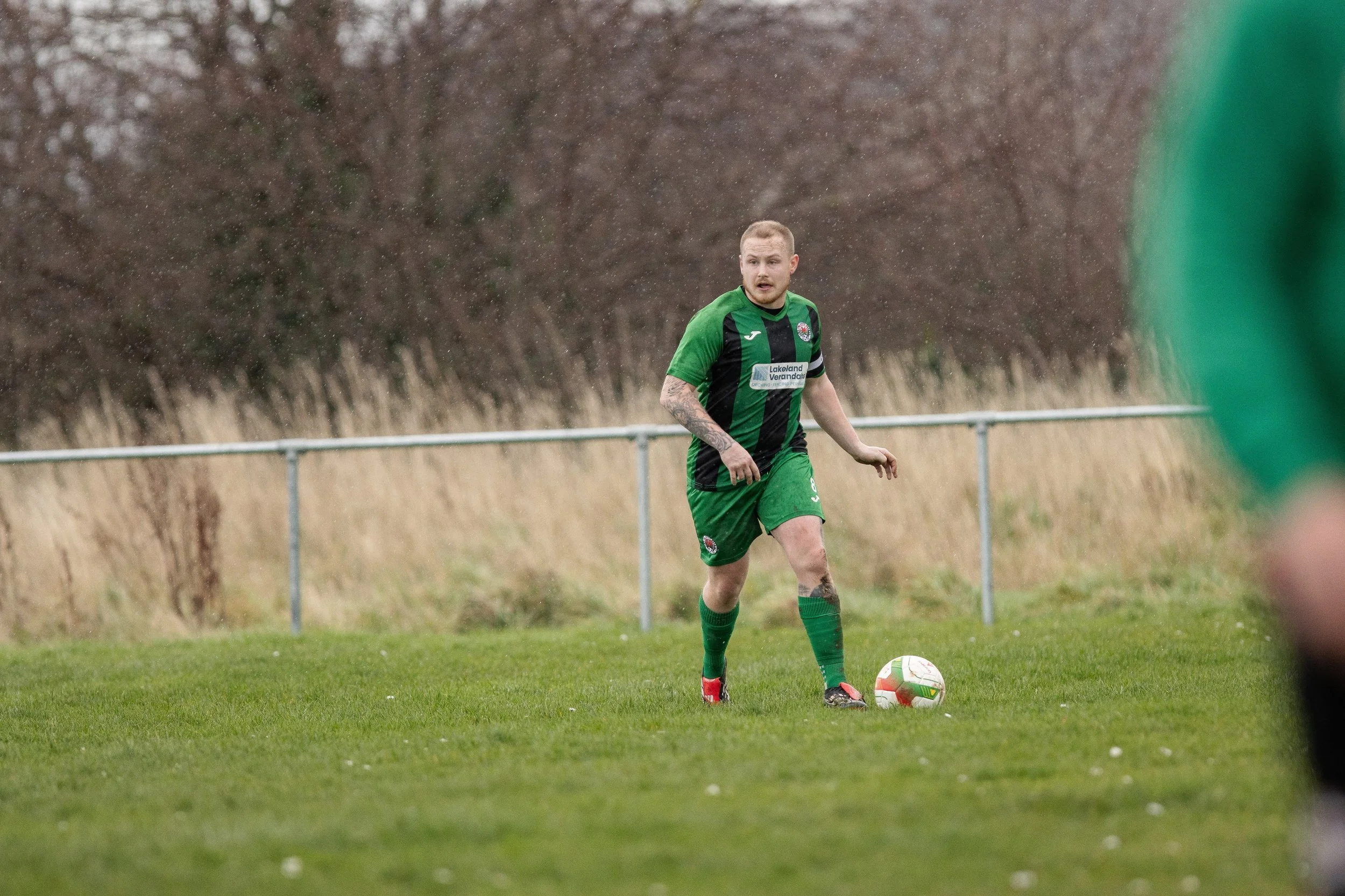 A soccer player in a green and black uniform standing on a grassy field with a soccer ball at his feet, during rainy weather.