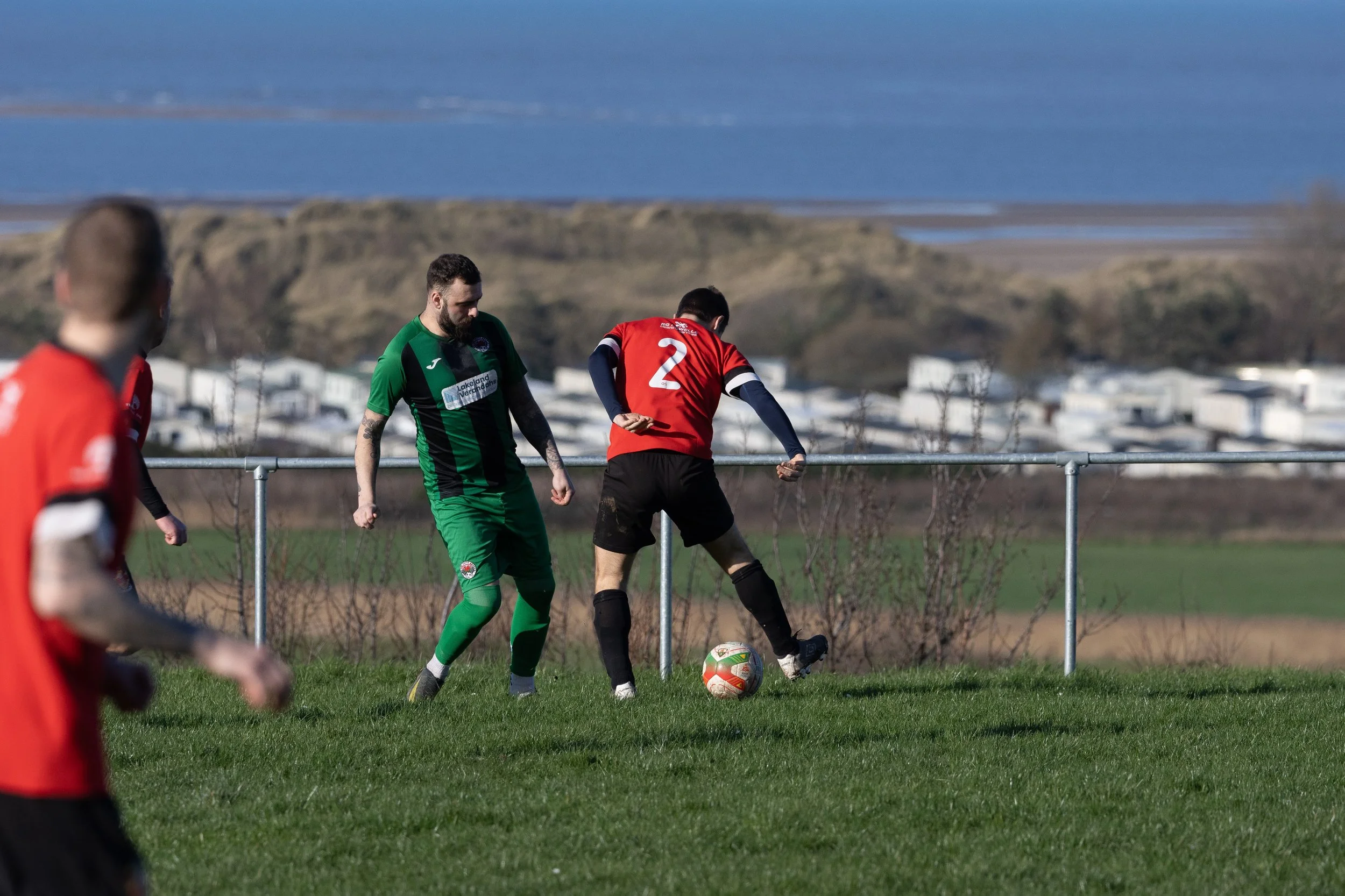 Soccer players in red and green uniforms competing for the ball on a grassy field with hills and water in the background.