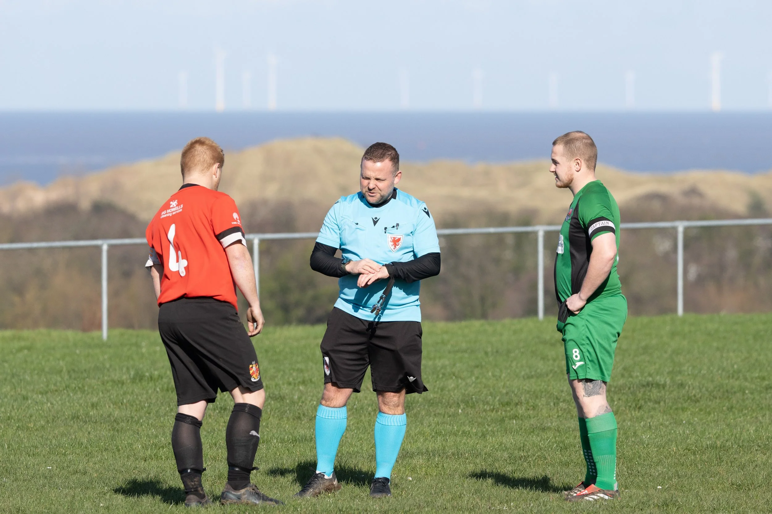Two soccer players and a referee standing on a grassy field, with hills and wind turbines in the background.