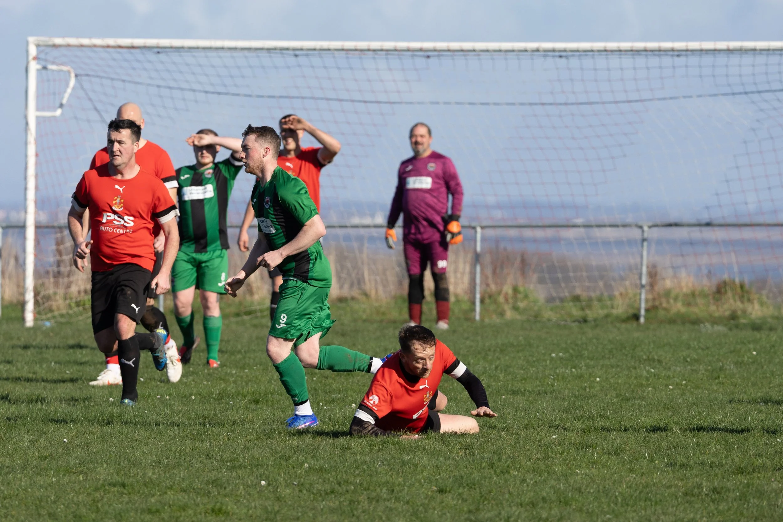 Soccer players on the field, one on the ground reaching for the ball, others standing near the goal, goalkeeper in purple in the background, during a match on a grassy field