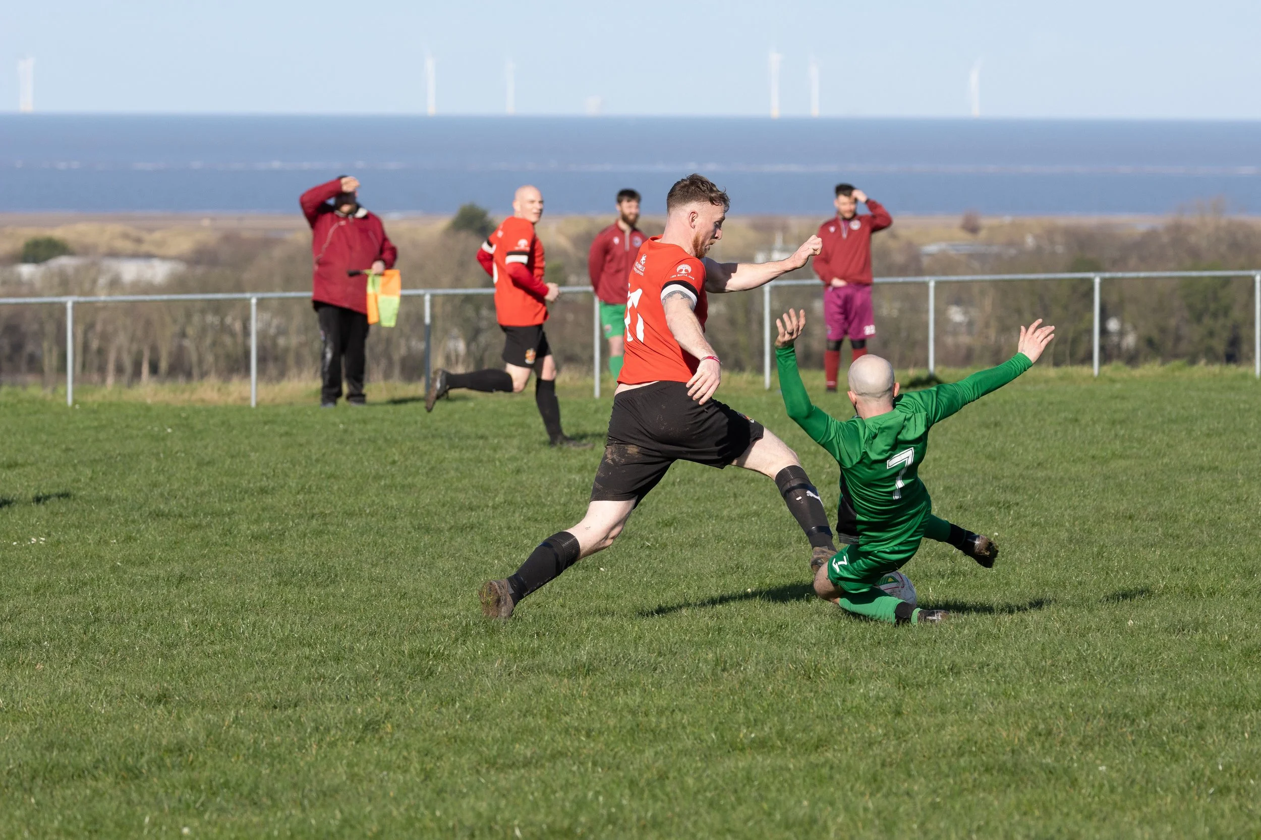 A soccer match showing an outfield player in a red jersey kicking the ball and a goalkeeper in a green jersey on the ground trying to block it on a grassy field with a scenic background.