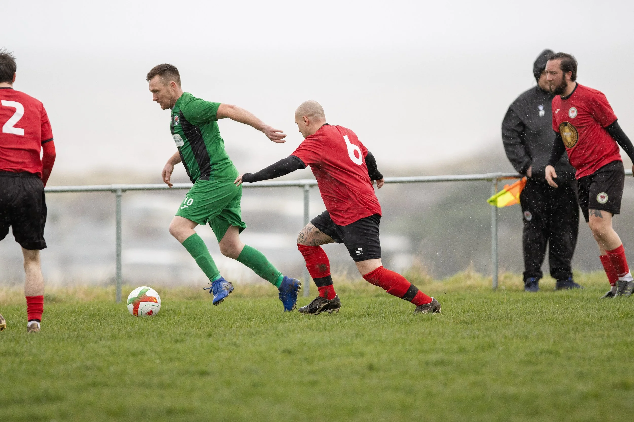 Soccer players in green and red jerseys competing for the ball on a grassy field on a rainy day, with one player in green dribbling the ball while a player in red attempts to tackle him.