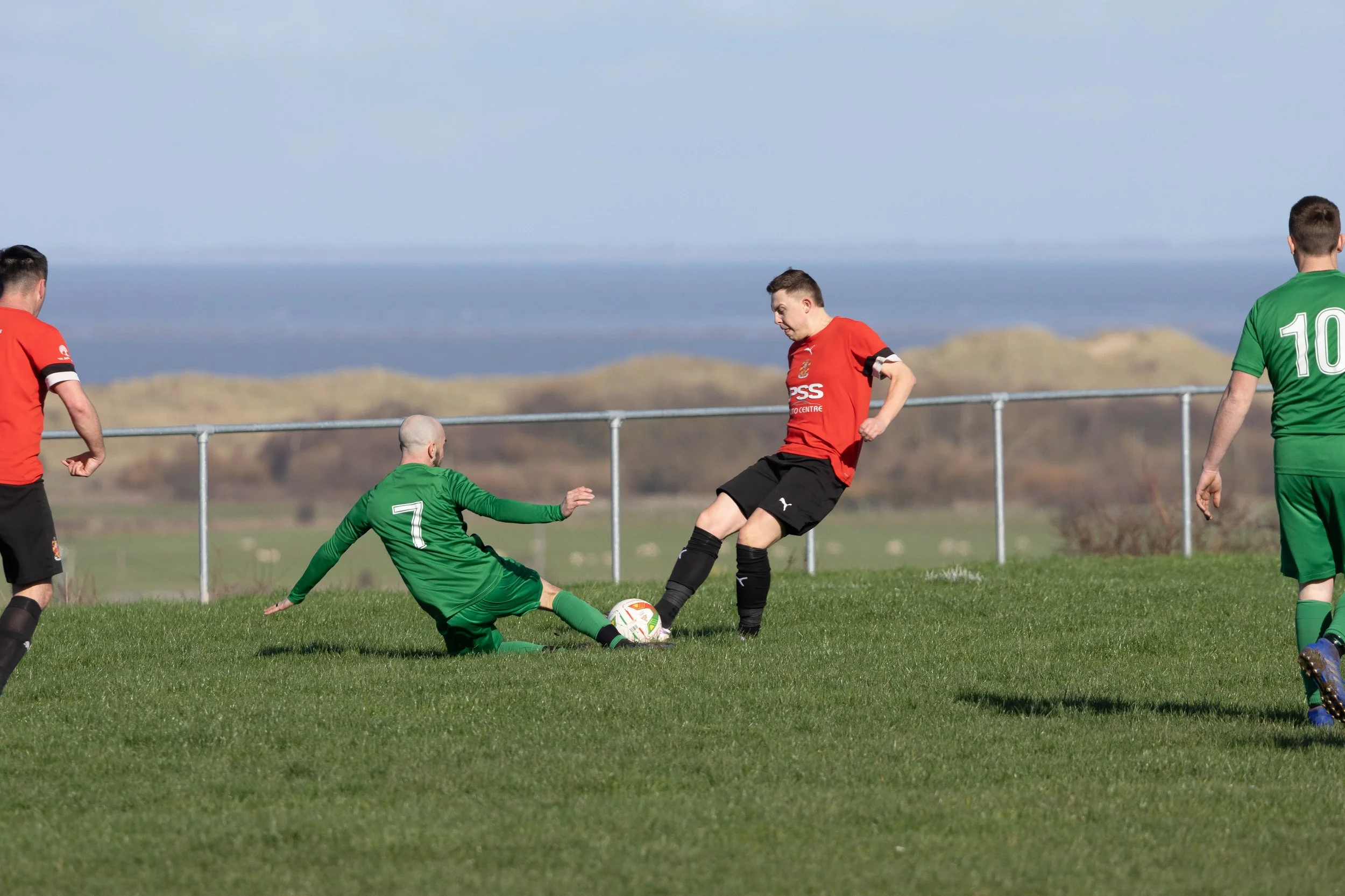 Soccer players in green and red uniforms competing for the ball on a grassy field, with a metal fence and hills in the background.