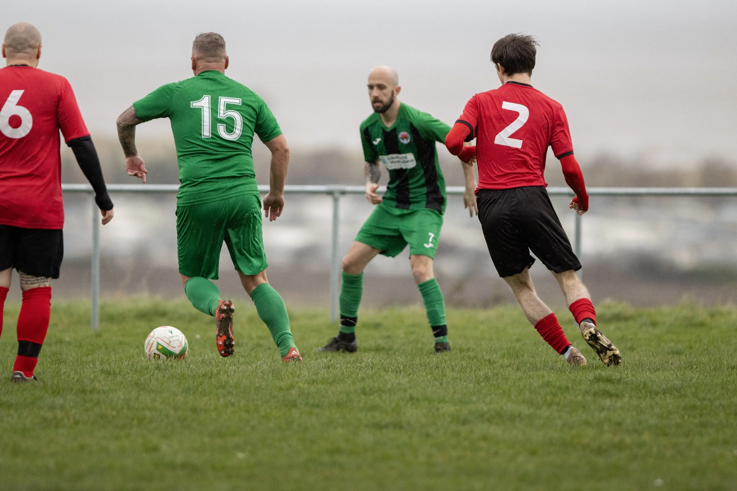 Soccer players in red and green jerseys compete for the ball on a grassy field with a railing and a foggy background.