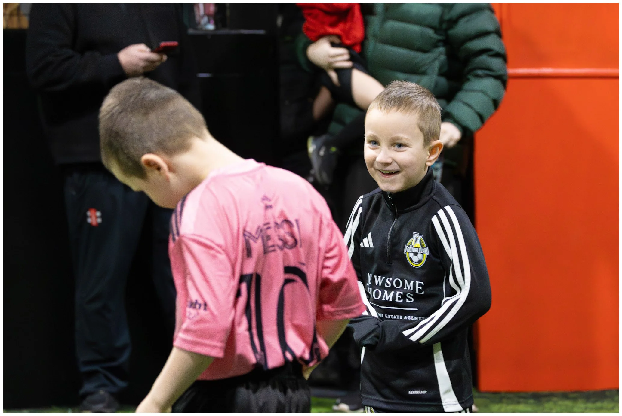 Two young boys are at a sports facility, with one boy smiling and holding the other boy's arm. The boy smiling is wearing a black sports jacket with white stripes and a logo, while the other boy is wearing a pink sports jersey with the number 10 on i