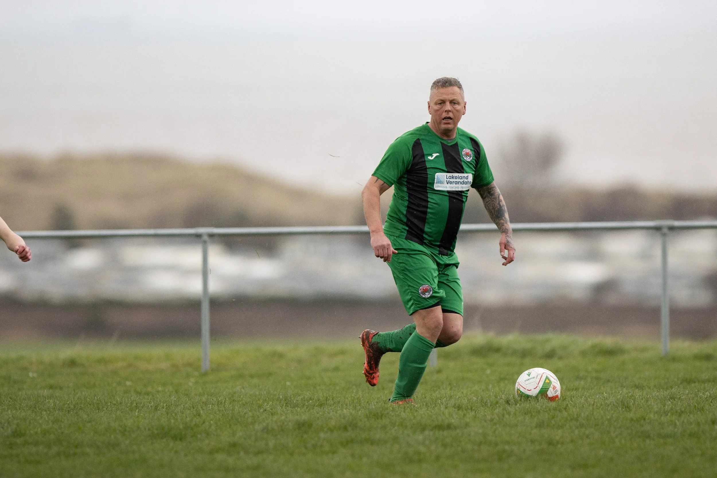 A man in a green and black soccer uniform running on a grass field with a soccer ball at his feet during an overcast day.