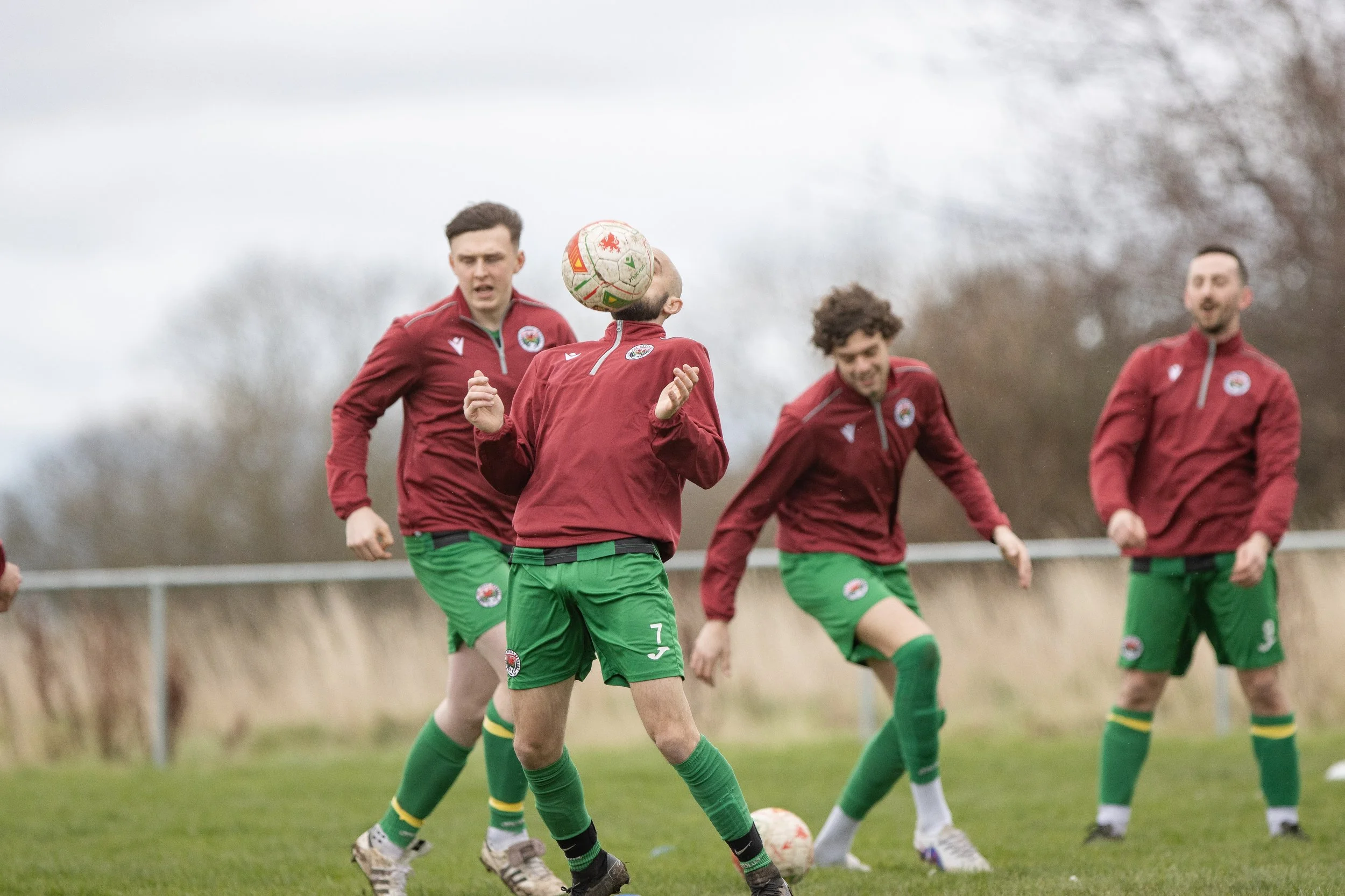 Group of four male soccer players in maroon jackets and green shorts practicing on a soccer field with a soccer ball.