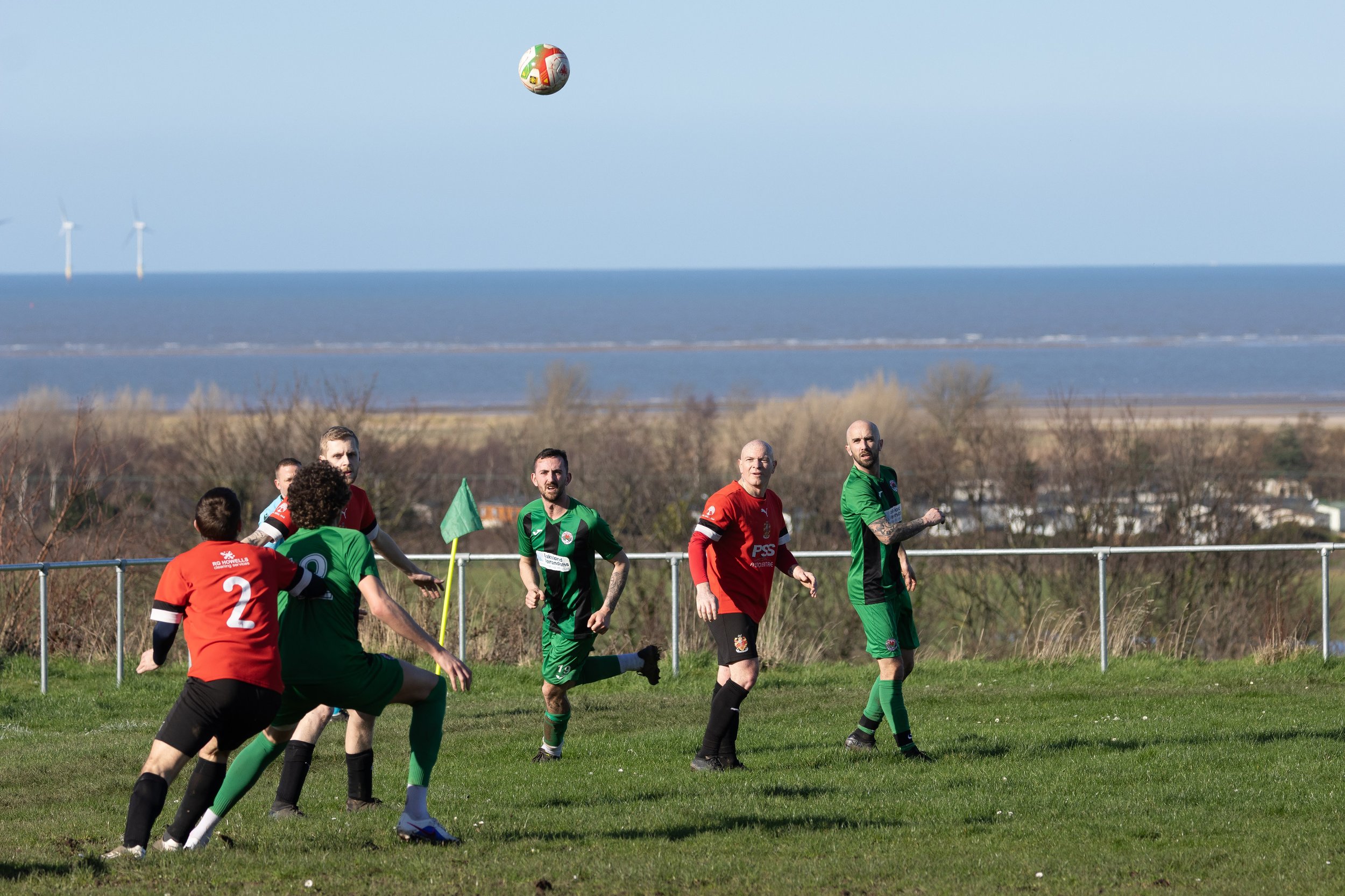 Soccer players in green and red uniforms playing on a grassy field near the coast, with wind turbines in the ocean in the background, under a clear sky.