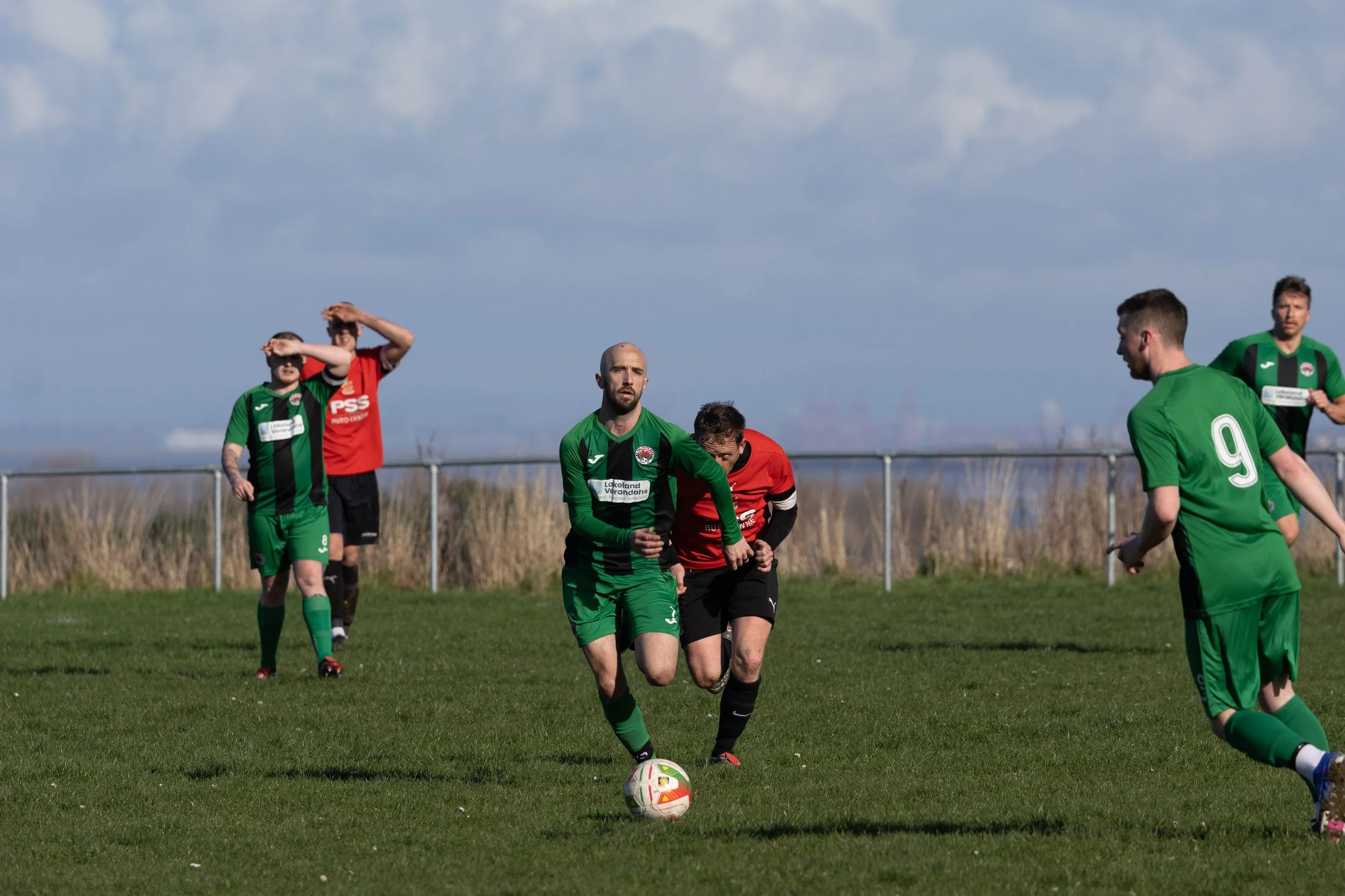 Soccer players on a grass field during a match, with some players running and one about to kick the ball, under a partly cloudy sky.