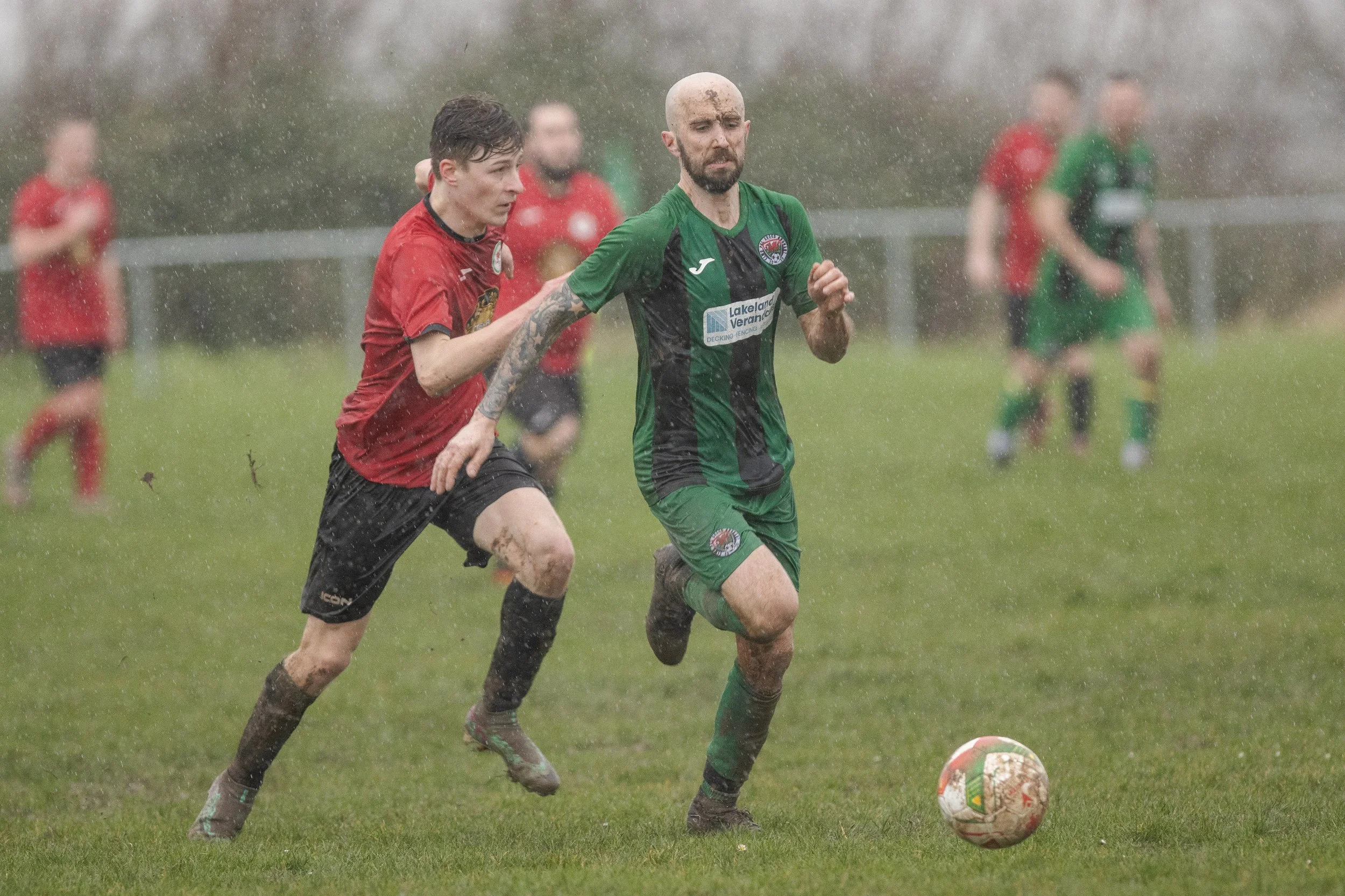Two soccer players running in the rain on a grassy field, one in a green and black kit and the other in a red and black kit, chasing a soccer ball while other players follow behind.