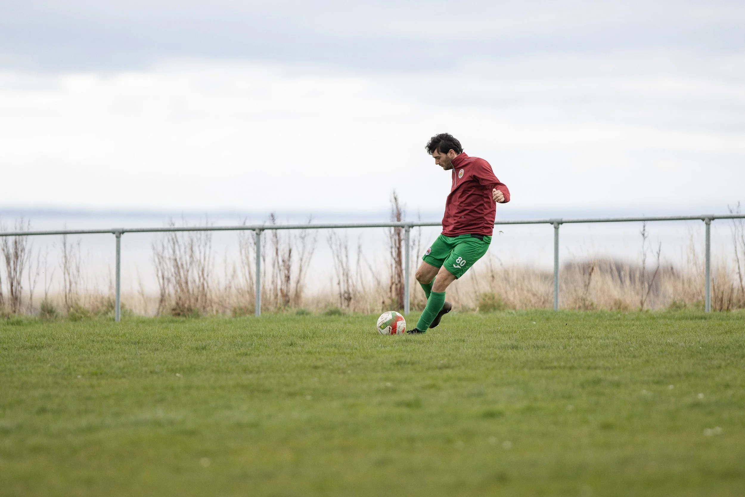 A man in a red shirt and green shorts playing soccer on a grassy field with a cloudy sky overhead.