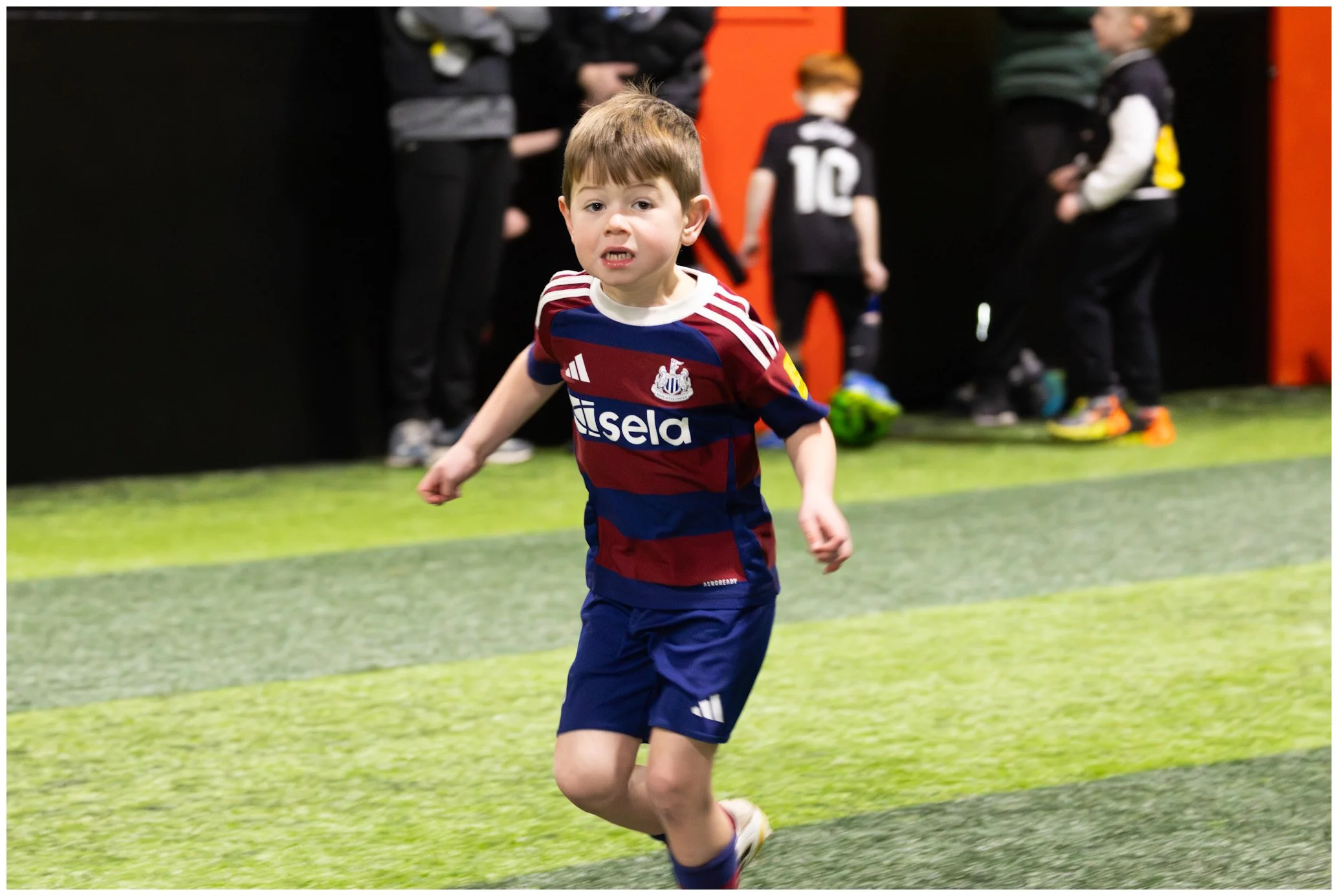 A young boy with brown hair running inside an indoor sports facility, wearing a maroon and blue striped soccer jersey and blue shorts, with a concerned or determined expression on his face. In the background, other children and adults are standing ne