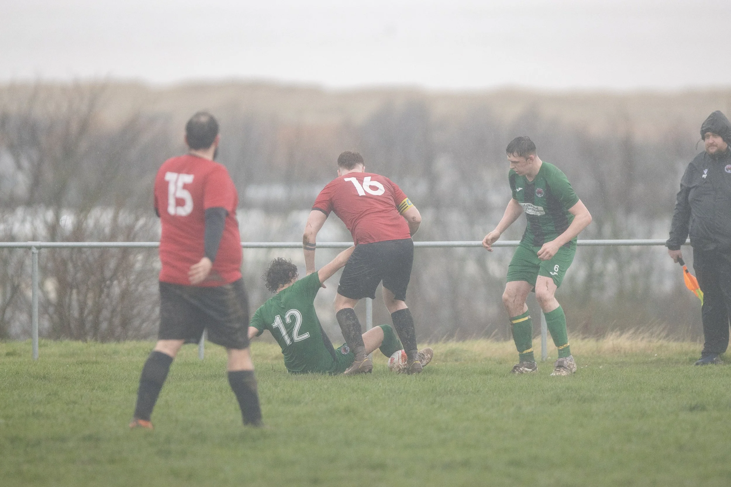 Soccer players in green and red uniforms competing for the ball on a grassy field, with a referee in black holding a yellow flag