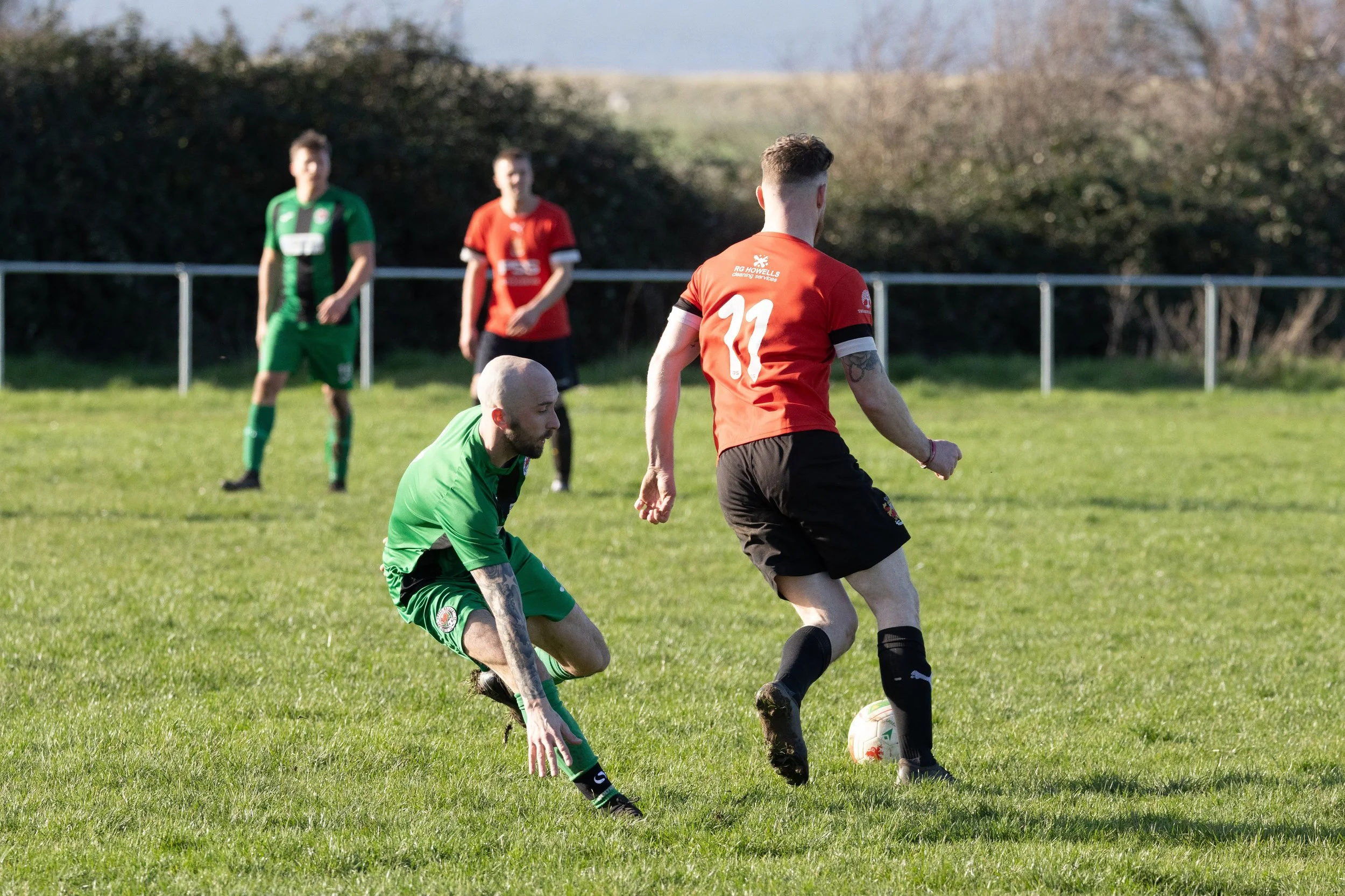 A soccer match taking place on a green field with four players visible. One player in a red shirt and black shorts is dribbling the ball, while another player in a green shirt and shorts is attempting to intercept or defend. Two additional players, a