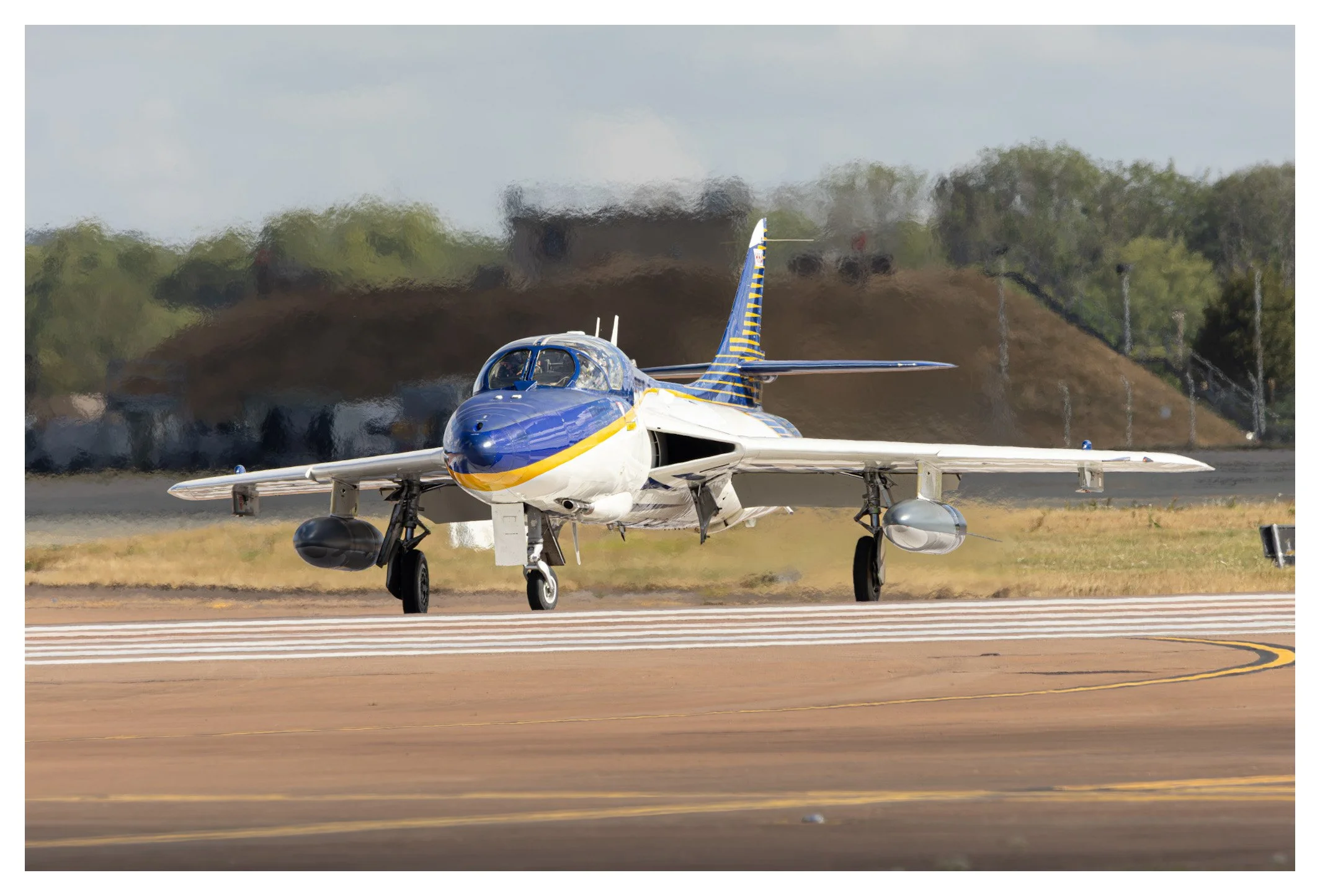 A blue and white fighter jet on a runway, with a blurred background of trees and airport structures.