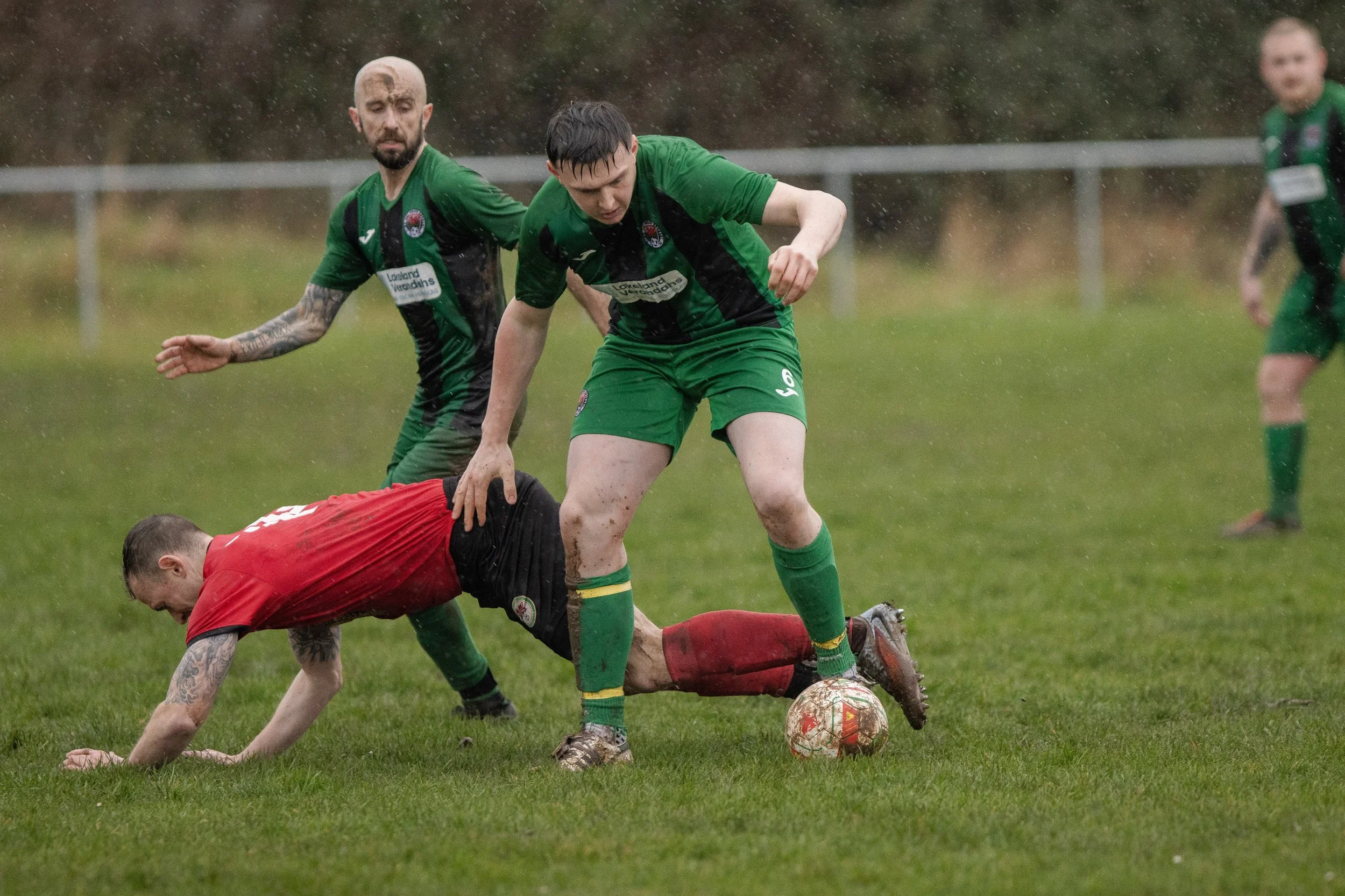 Soccer players in green and red uniforms play in the rain on a grassy field, with one player in red on the ground and another in green controlling the ball.