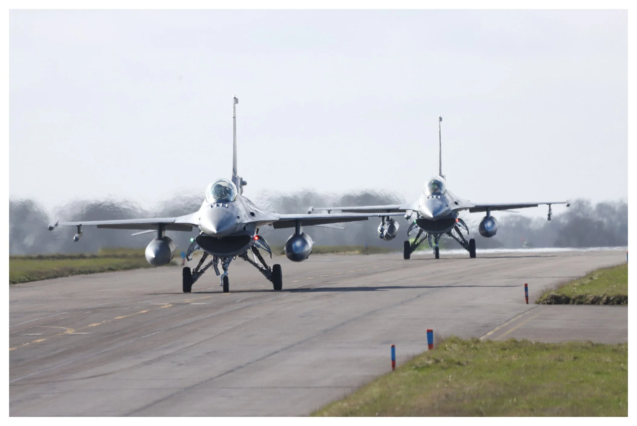 Two fighter jets taxiing on a runway with grass on the sides, preparing for takeoff or landing.