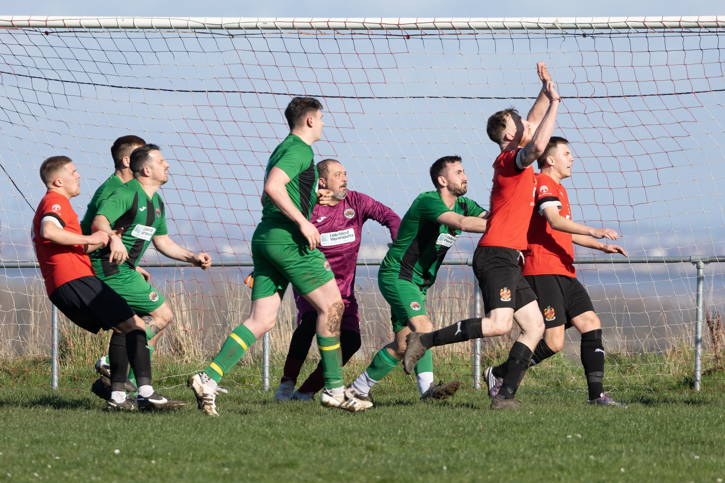 Soccer players in green and red jerseys contest a header near the goal during a match on a grassy field with a blue sky background.