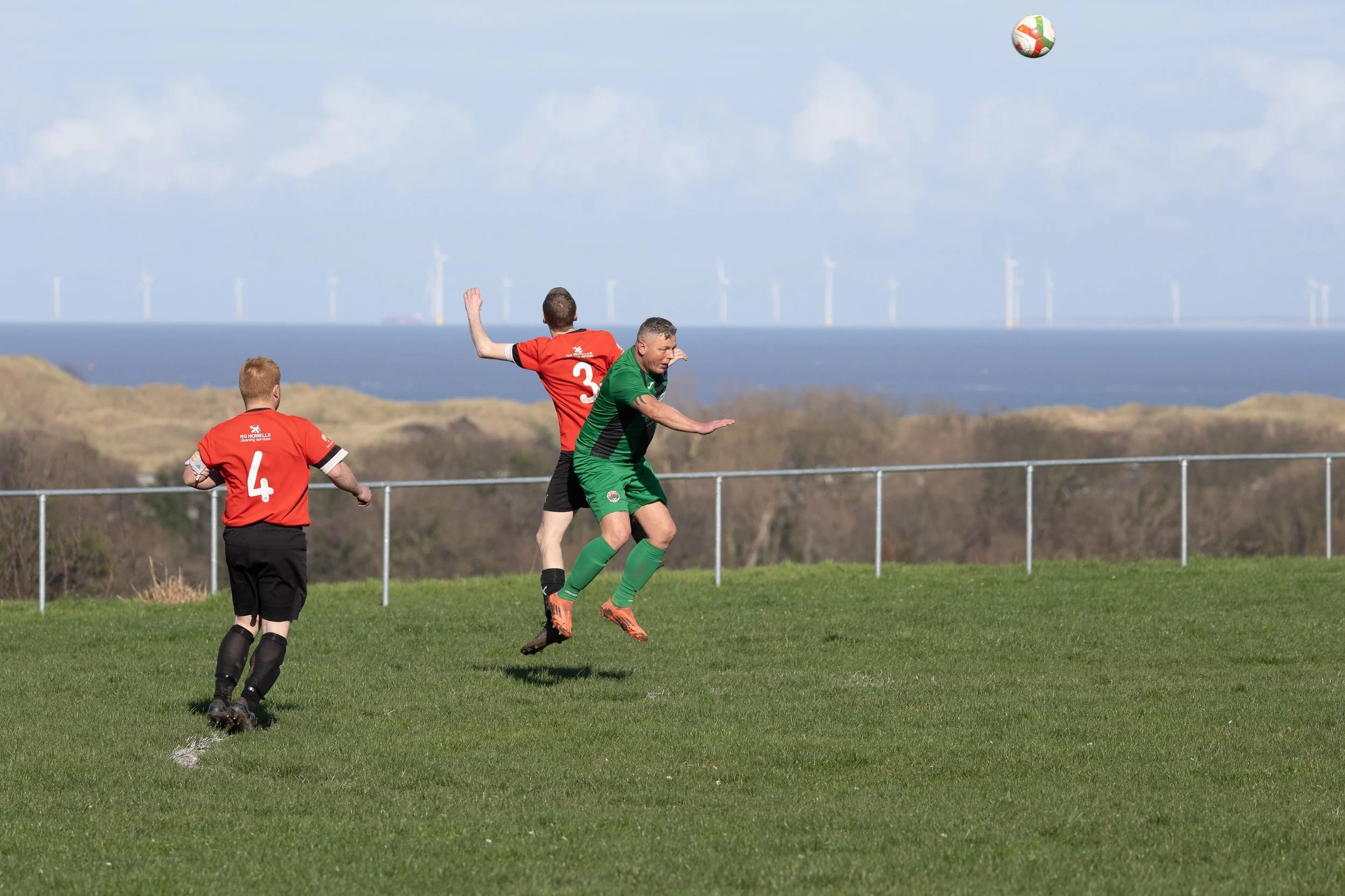 Three soccer players in red and green uniforms competing for the ball outdoors with wind turbines in the background under a cloudy sky.