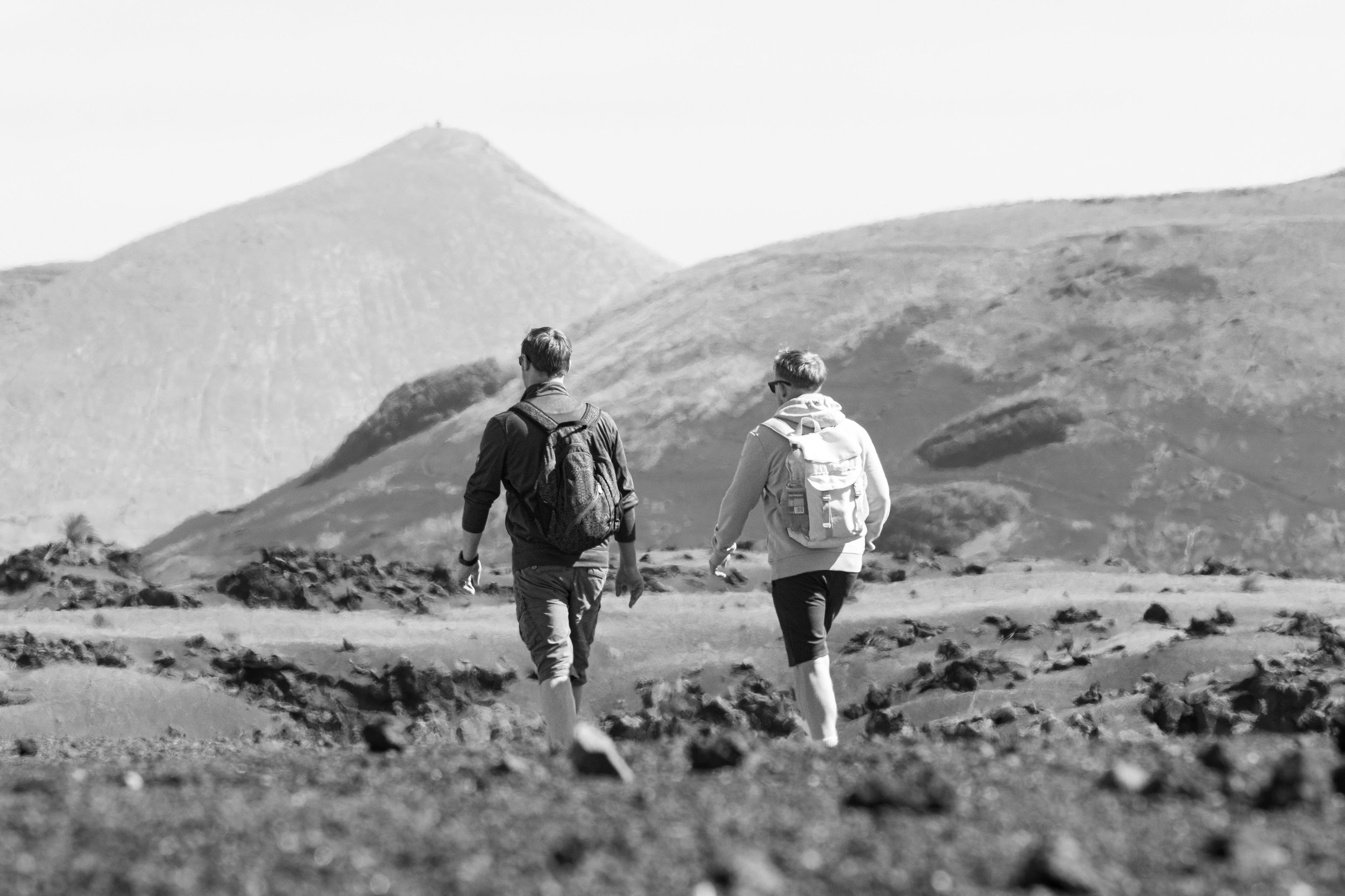 Two hikers with backpacks walk across a rocky plain with hills in the background.
