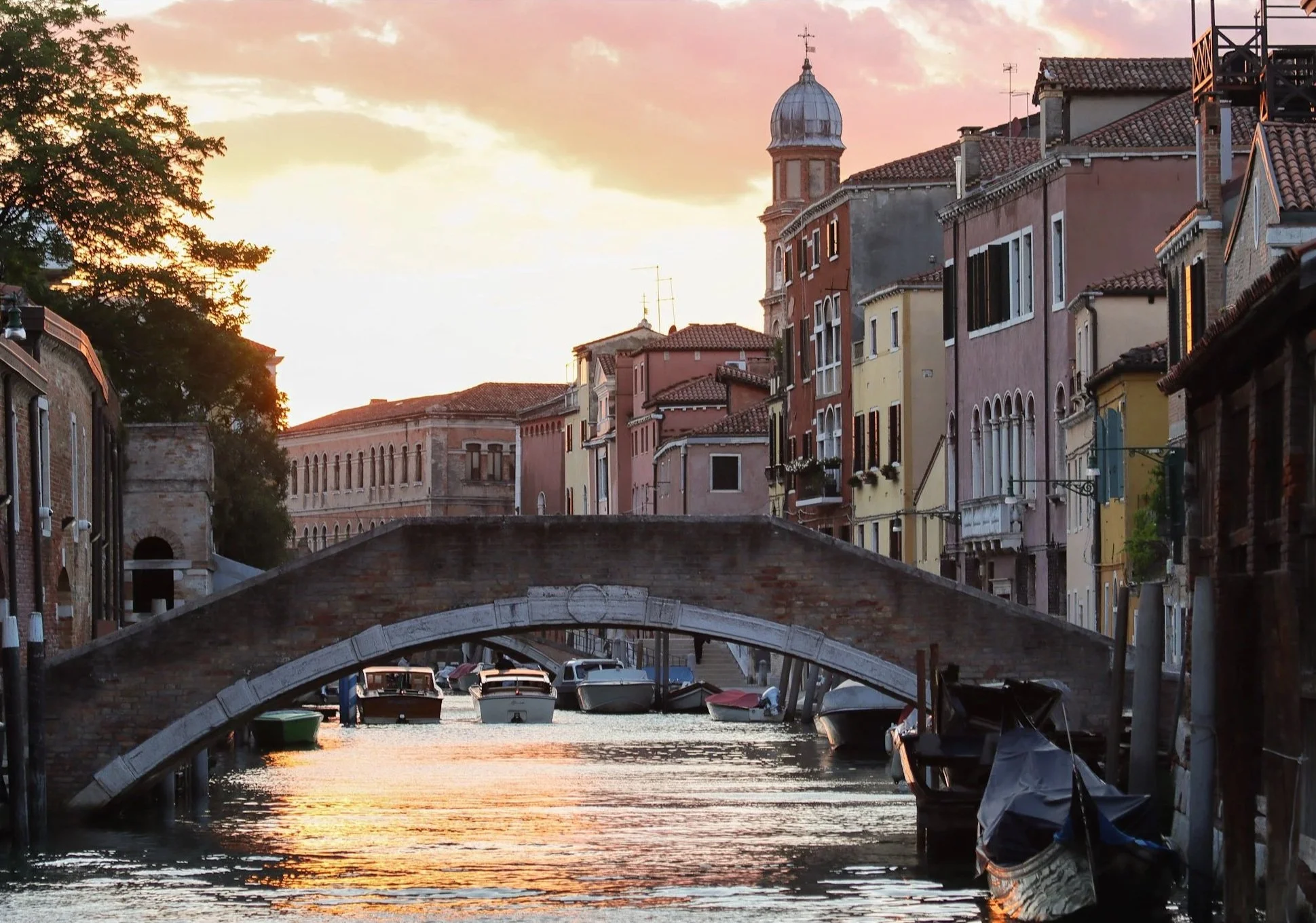Sunset over a canal in Venice, Italy with colorful buildings, a small stone bridge, and boats docked along the water.