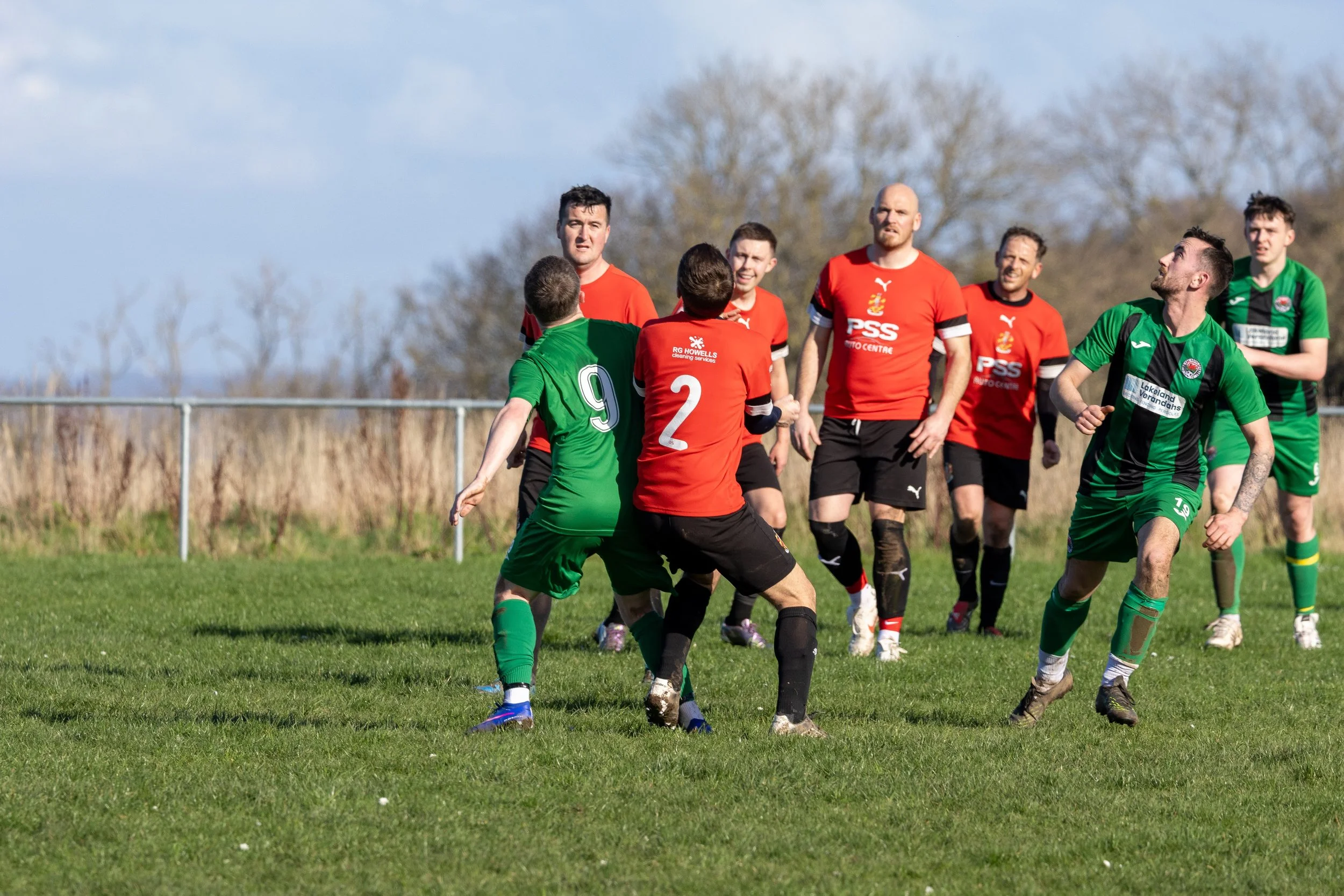 Soccer players on the field during a match, with some players wearing green and others wearing red jerseys. The players are engaged and positioned on a grassy field under a partly cloudy sky.