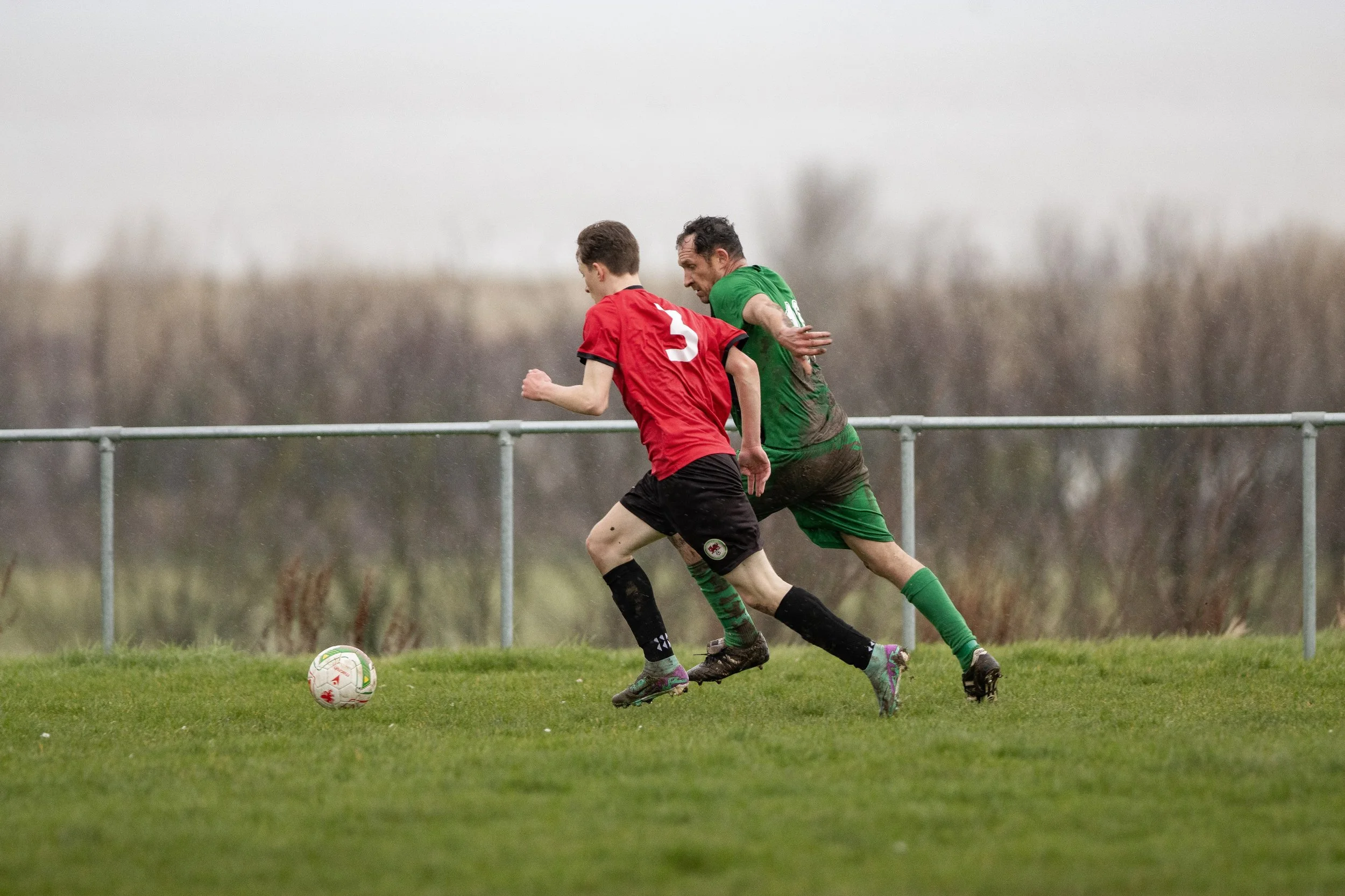 Two male soccer players in red and green uniforms running after a soccer ball on a grassy field, with a metal railing and leafless trees in the background, under cloudy skies.