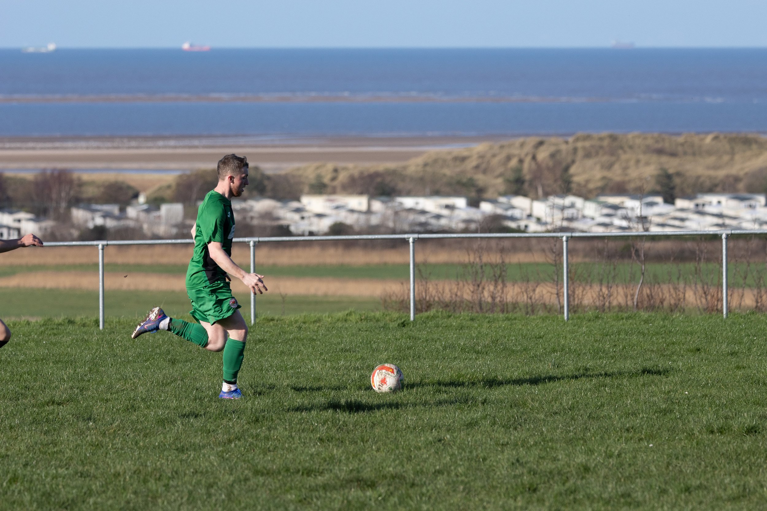 A soccer player in a green uniform running on a grassy field with a white and orange soccer ball, with a seaside landscape and small white buildings in the background.