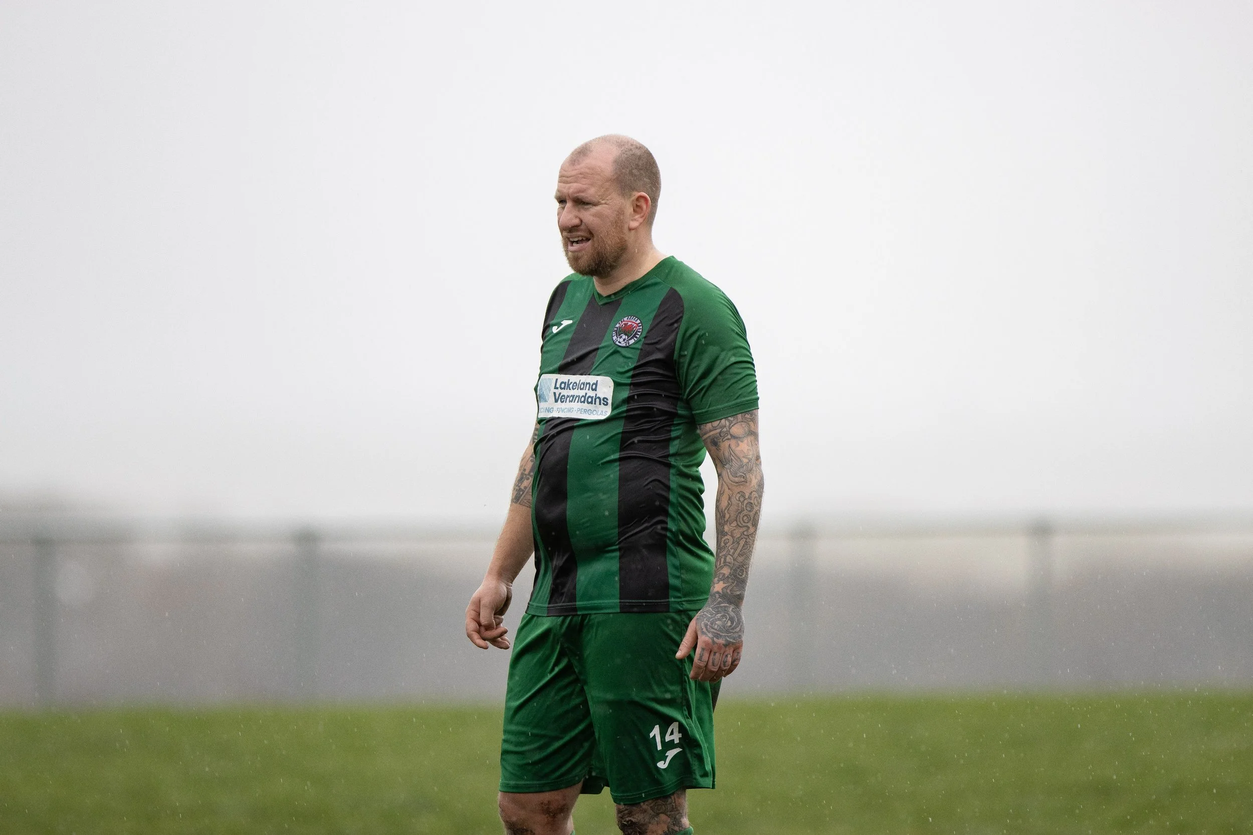 A male soccer player with tattoos wearing a green and black jersey stands on a field in rainy weather.