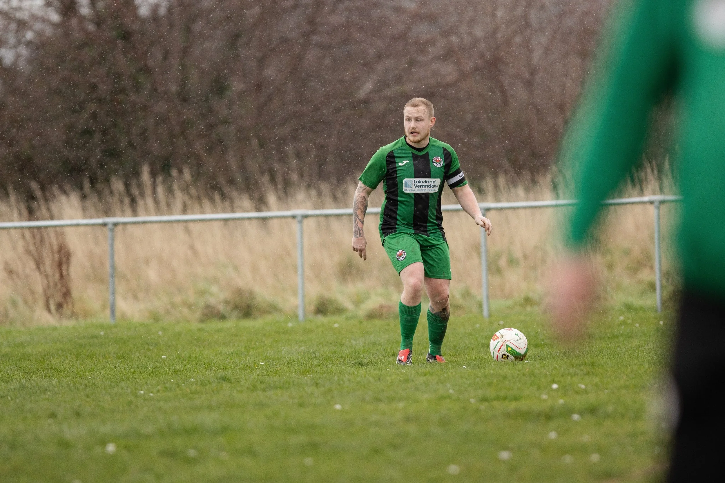 A soccer player with tattoos in green and black uniform preparing to kick a ball on a grassy field with a fence and trees in the background.