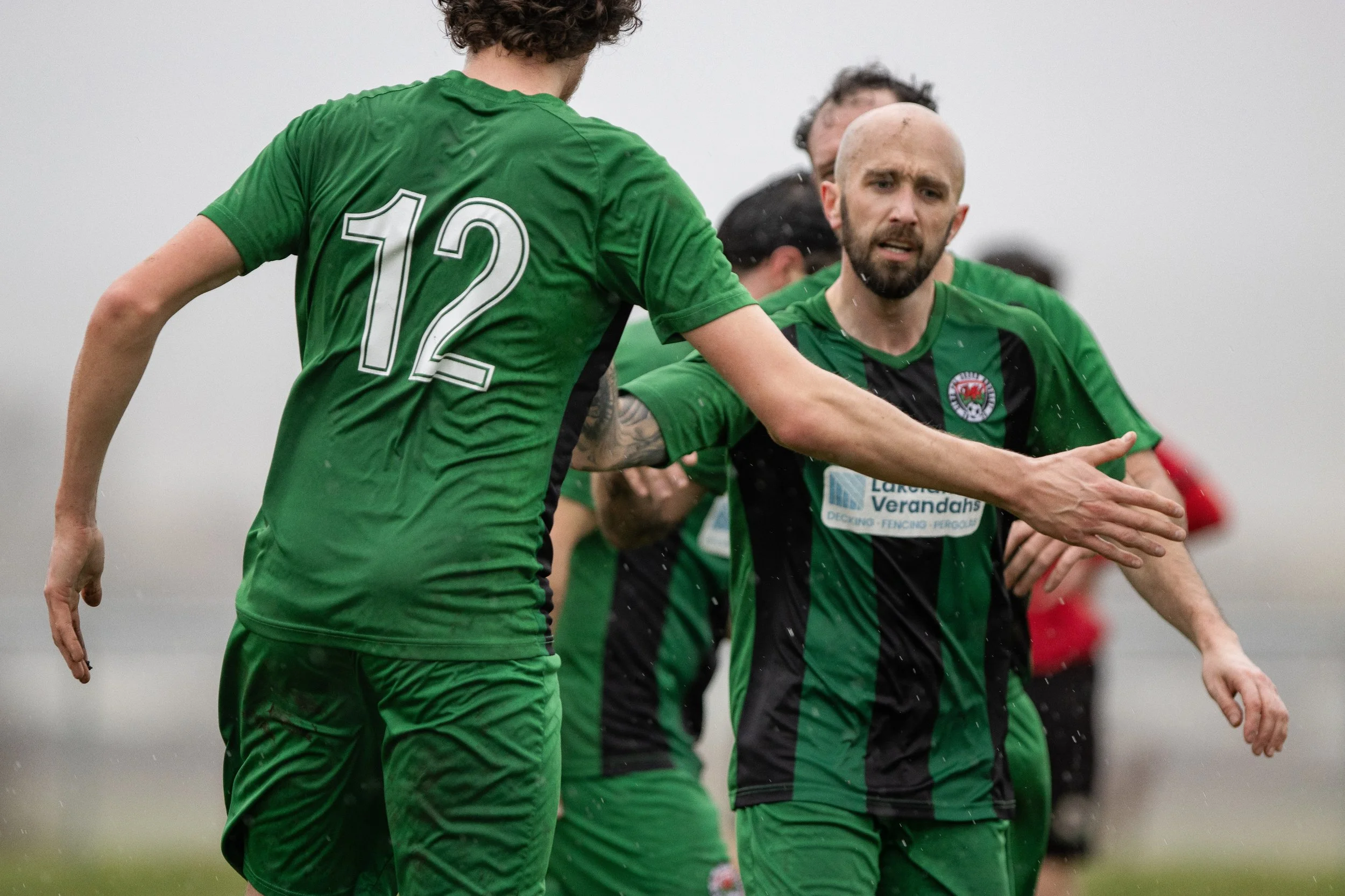 Soccer players in green and black uniforms celebrating on the field, one player with number 12 on his jersey.