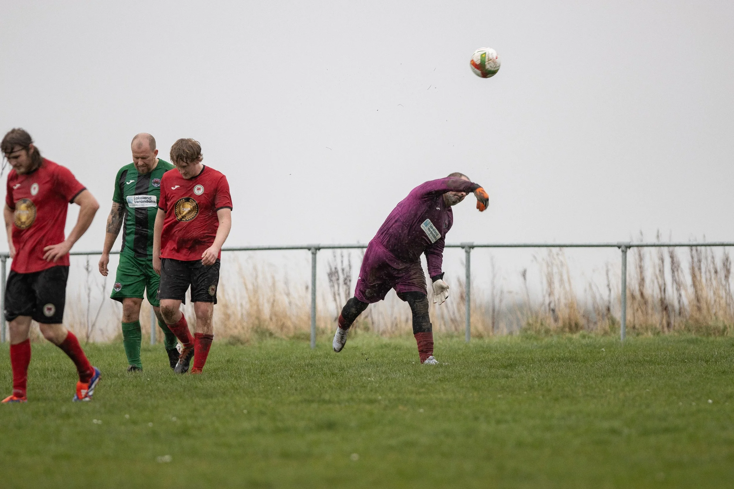 Soccer players on the field; one goalkeeper in purple uniform is preparing to catch or kick the ball, others are walking or standing, on a grassy field with a gray overcast sky and tall grass in the background.