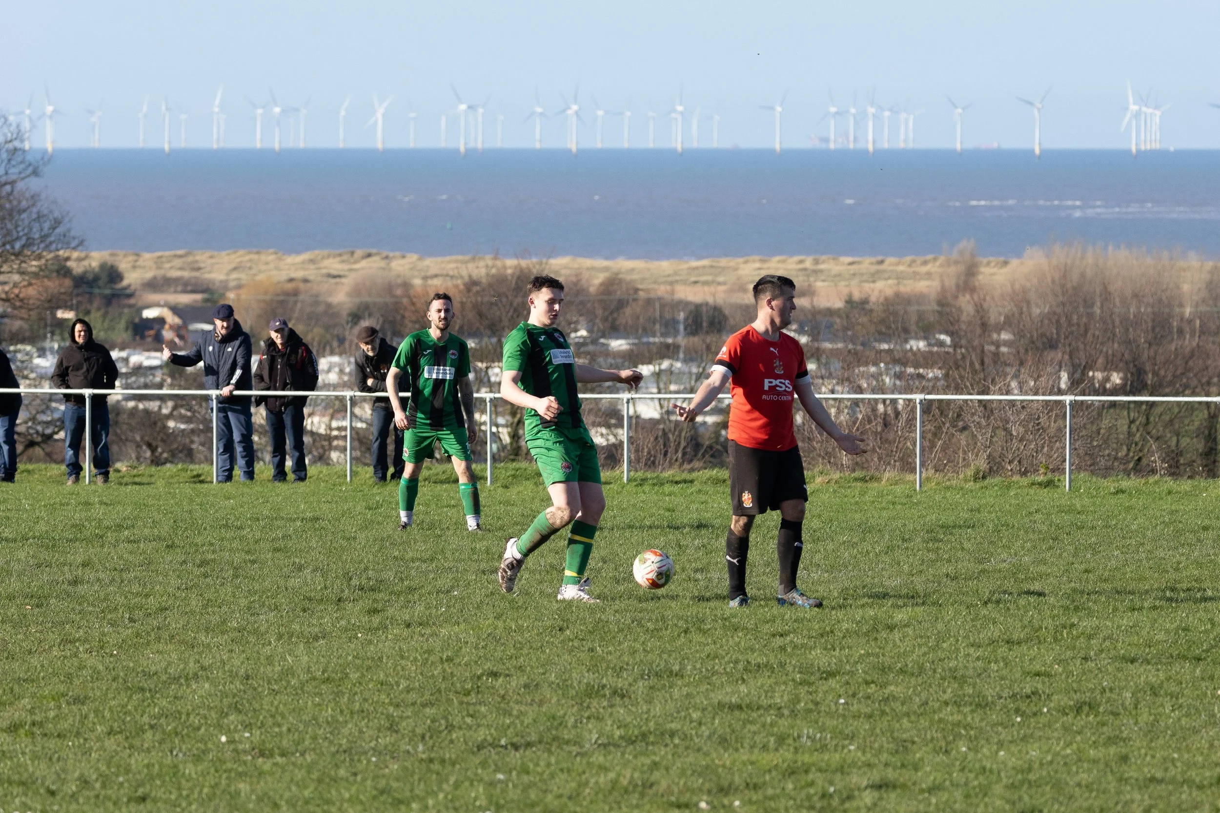 Soccer players on the field with one about to kick the ball, spectators watching from the sidelines, with wind turbines in the background under a clear sky.