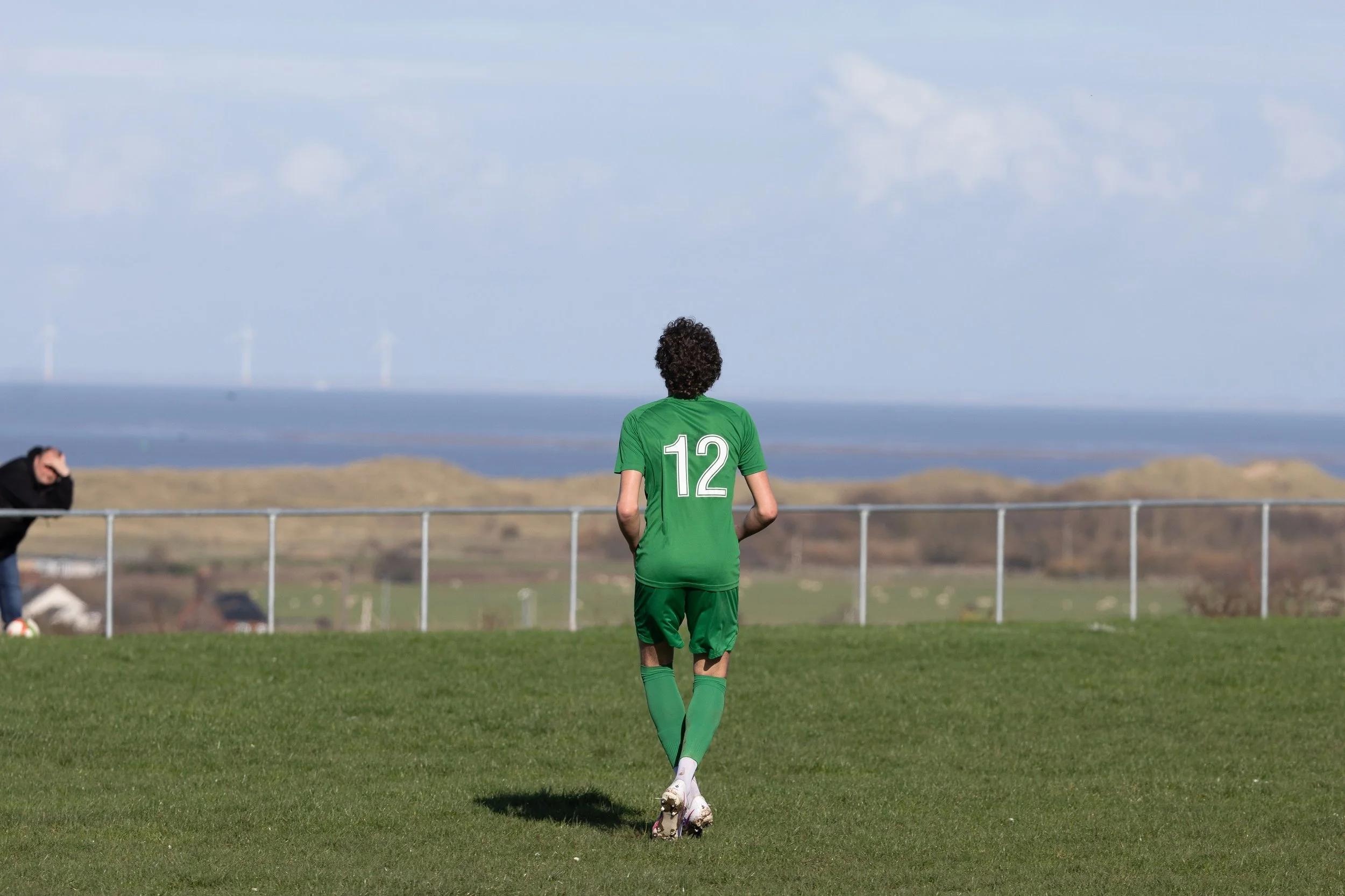 A soccer player in a green uniform with the number 12 on the back, walking on a grassy field with a distant landscape and blue sky in the background.
