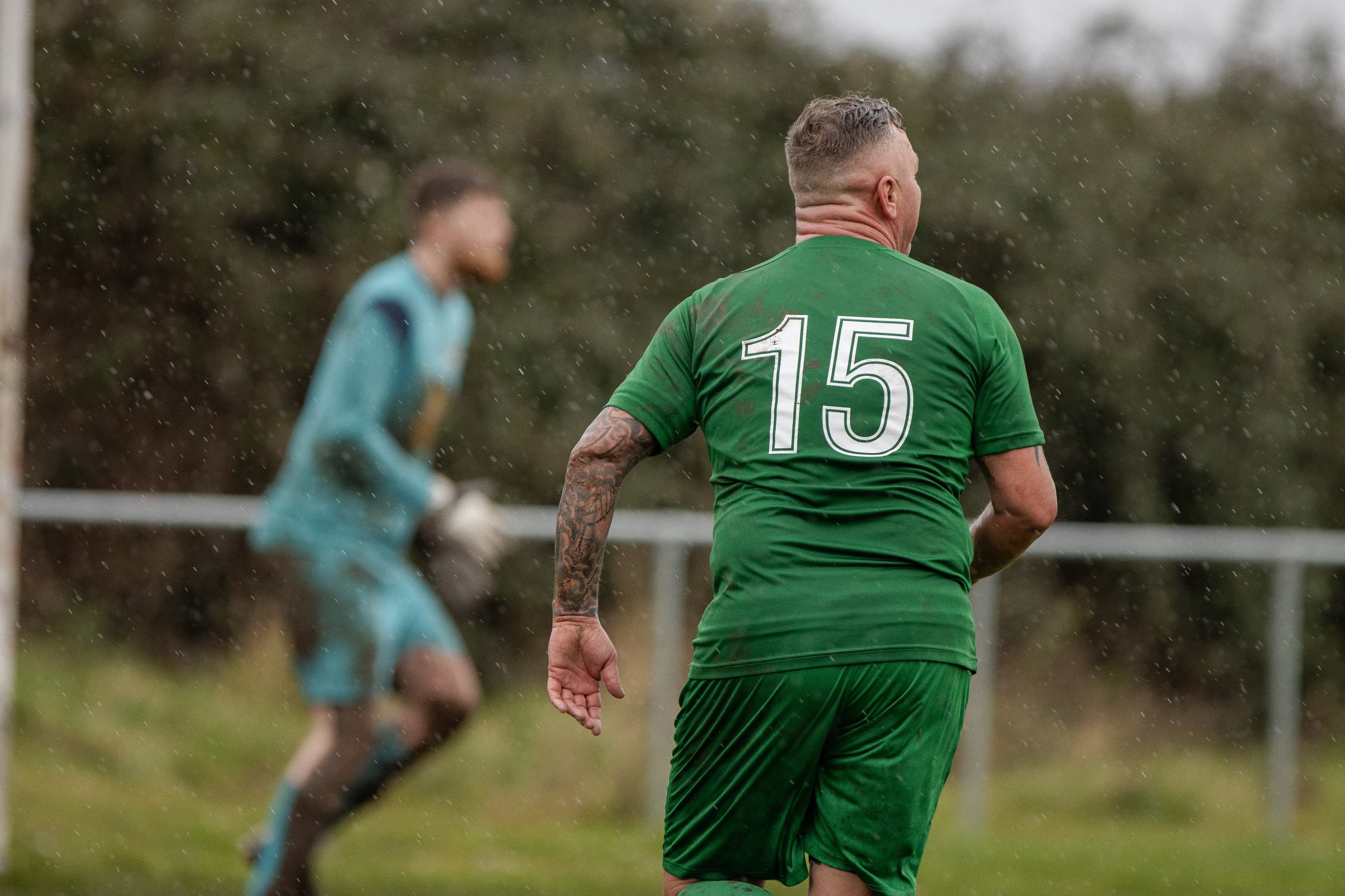 A soccer player in a green uniform with the number 15 on the back runs on the field, with a goalie in a teal uniform and gloves visible in the background, during a rainy day.