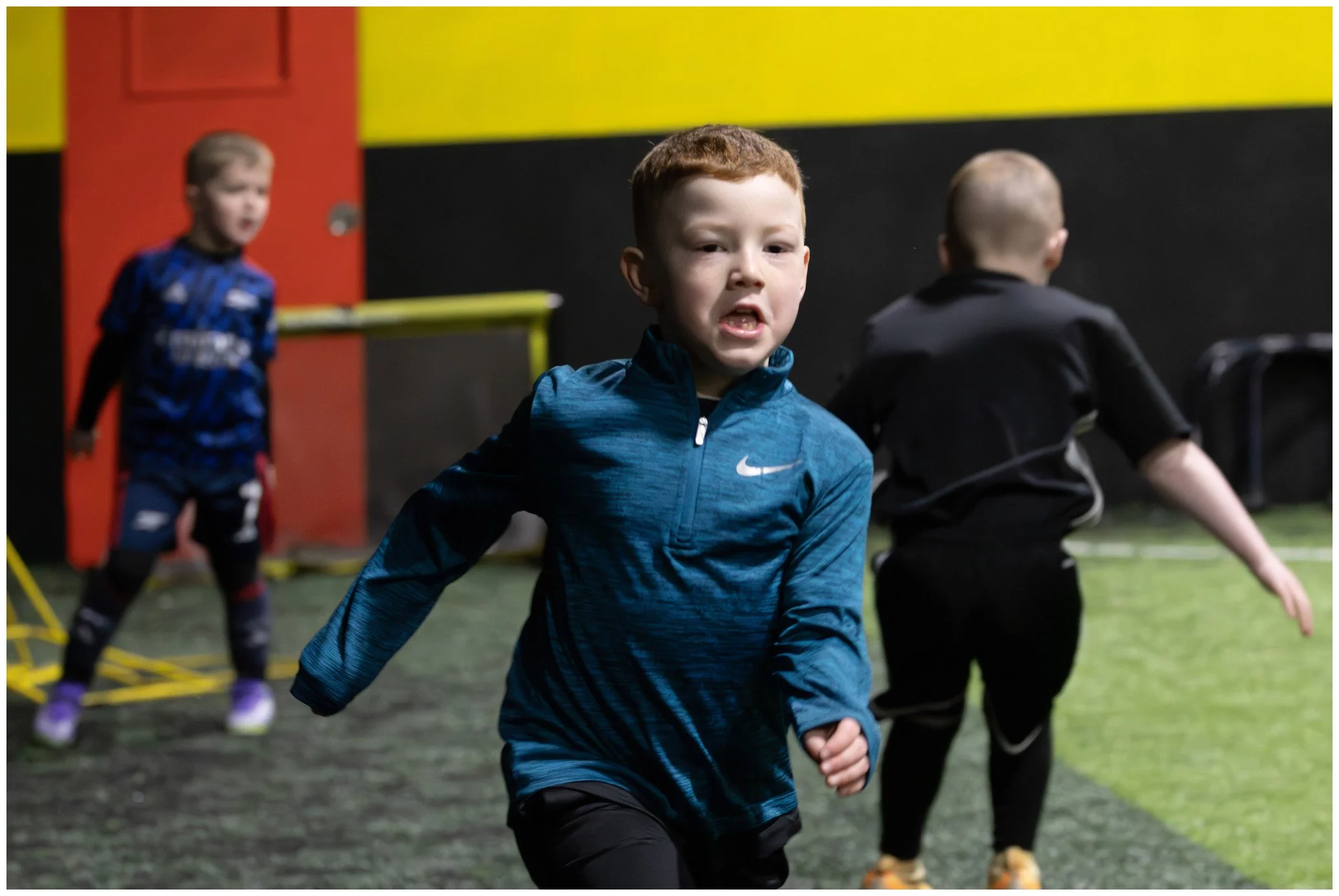 Young boys playing indoor soccer, with one in the foreground wearing a blue Nike jacket, running towards the camera, and two others in the background on a turf field.