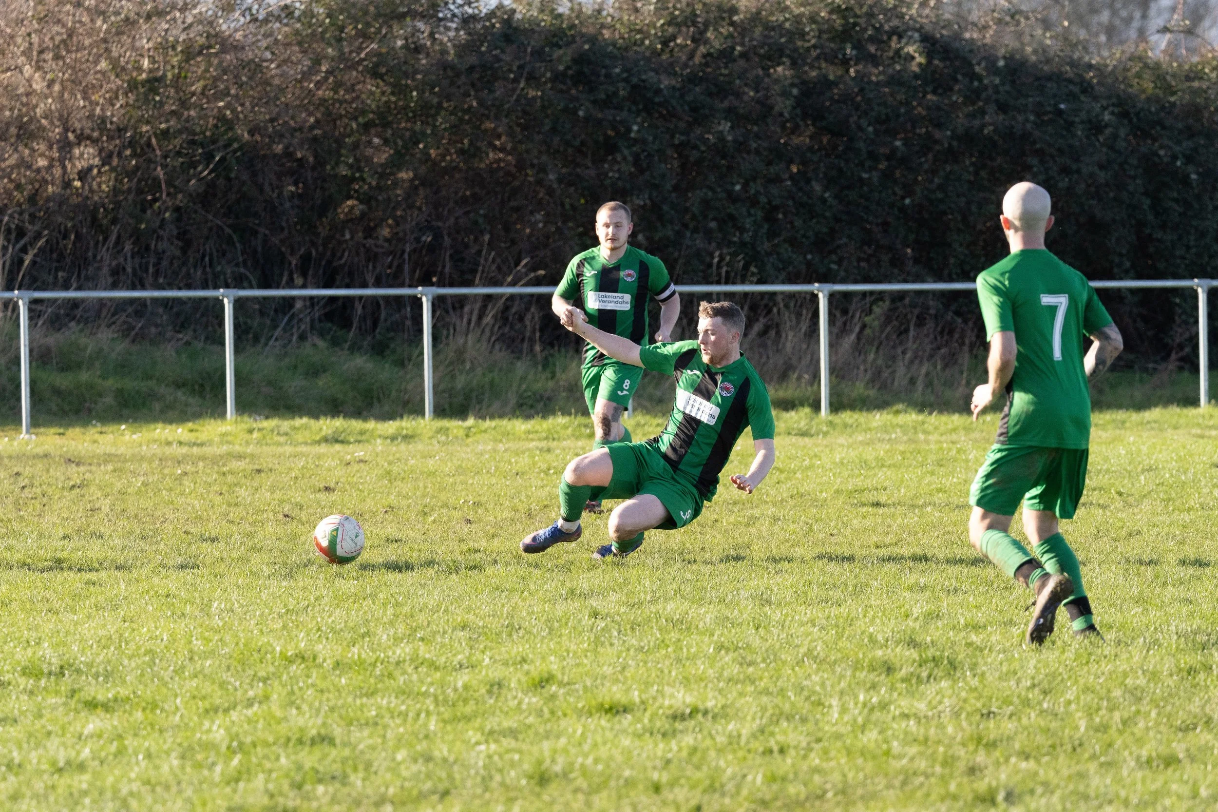 Three soccer players in green uniforms on the field during a game. One player is sliding to kick the ball while the others observe.