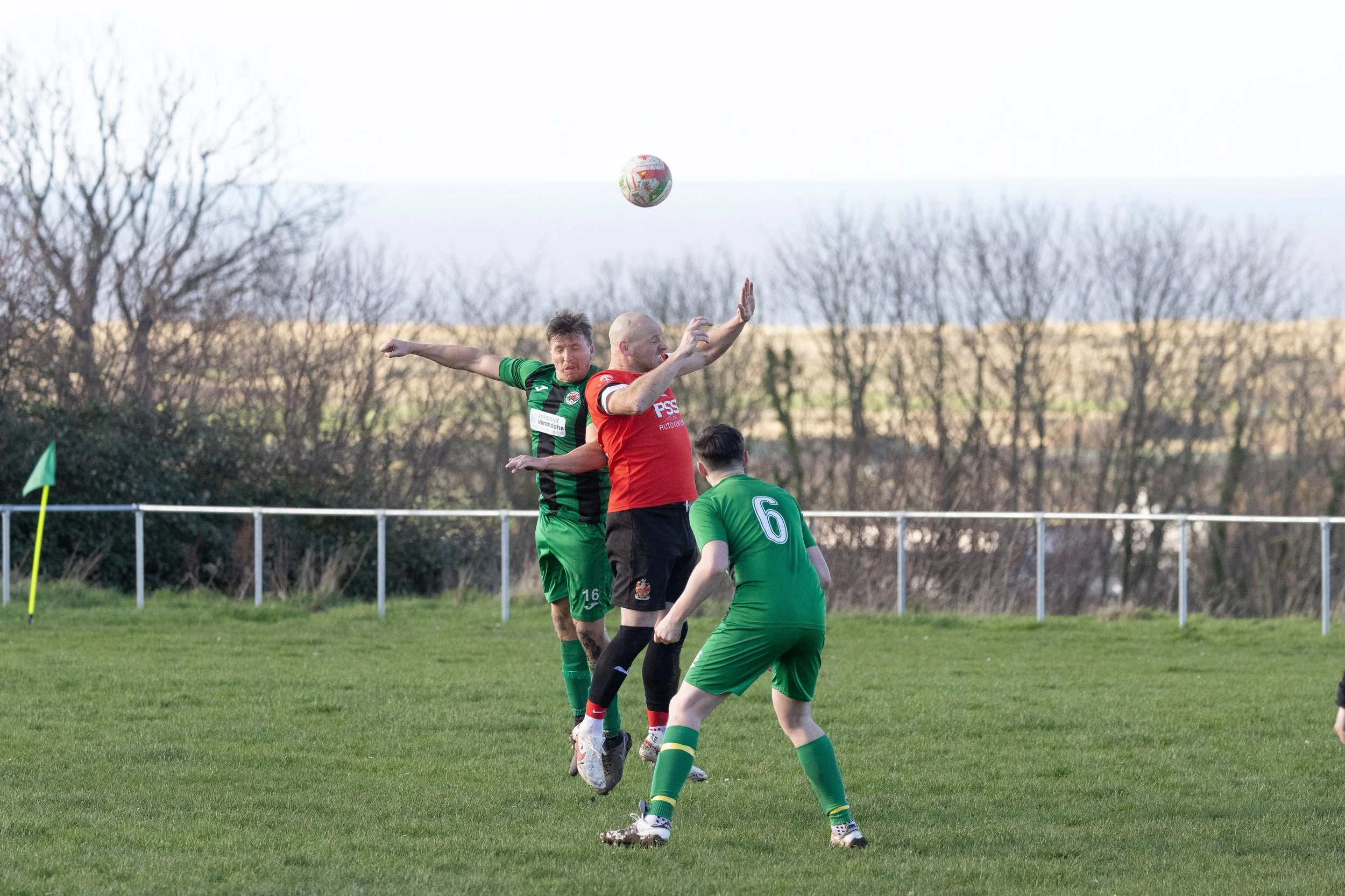 Three soccer players and a referee during a match. Two players in green uniforms are jumping to head the ball while the referee, in a red shirt, is nearby. The game is played on a grassy field with a fence and trees in the background.
