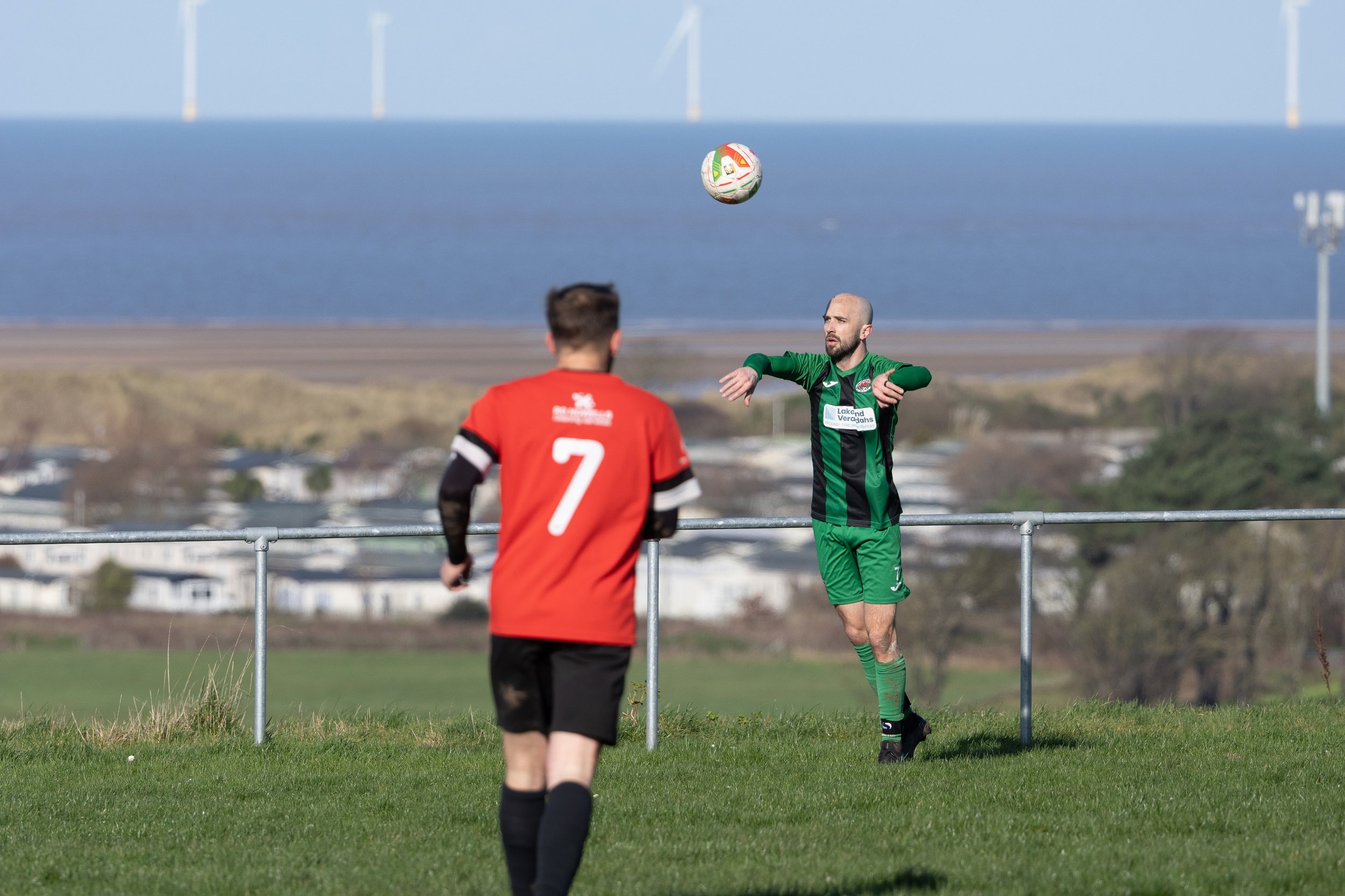 A soccer match with two players on a grassy field near a metal railing, one in a red jersey with the number 7, facing another in a green and black uniform who is preparing to kick the ball. In the background, there is a body of water and wind turbine