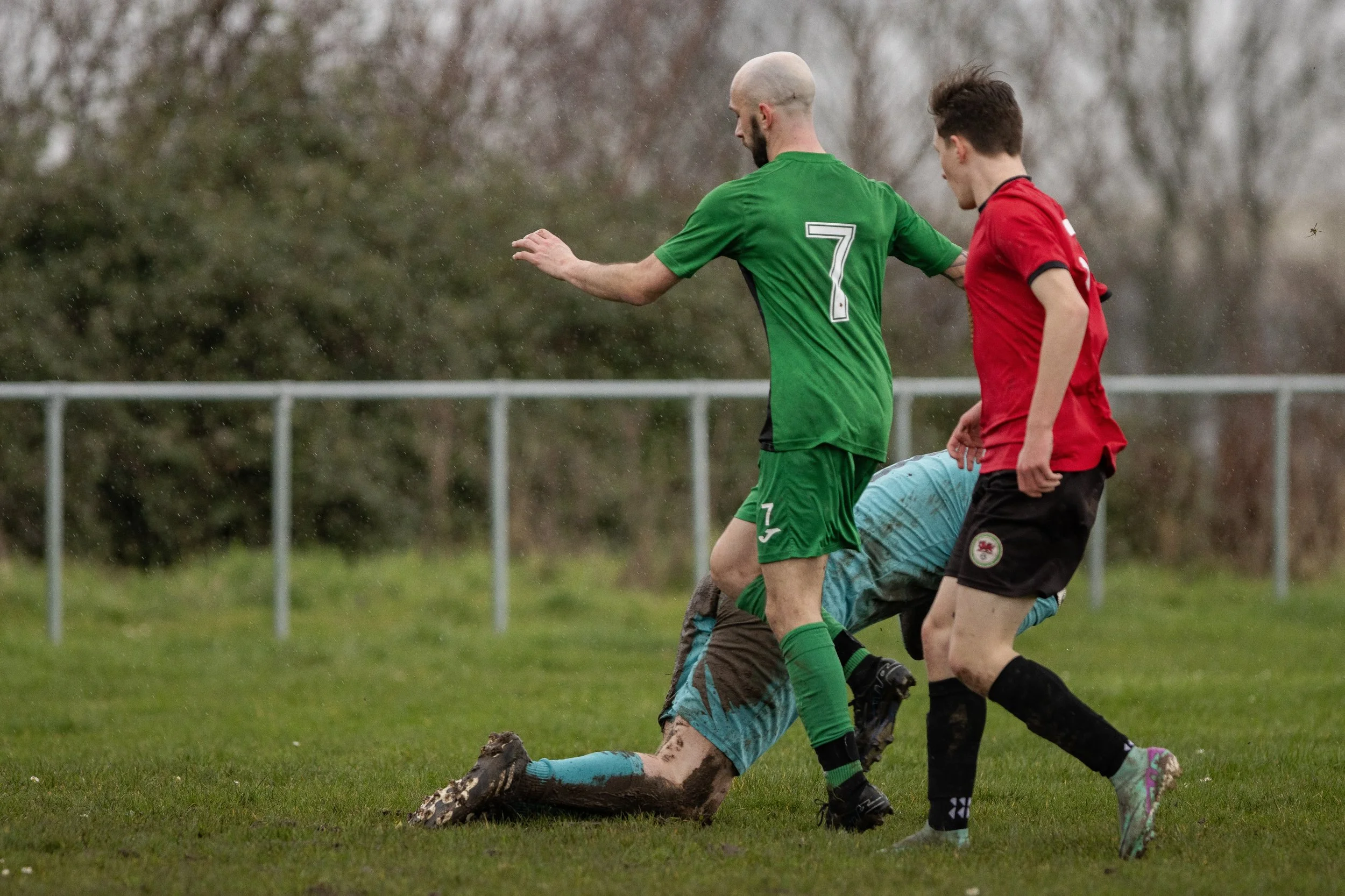 Soccer players during a match on a muddy field, with one player in a green uniform holding another player in a blue uniform on the ground, and a third player in a red jersey standing nearby.